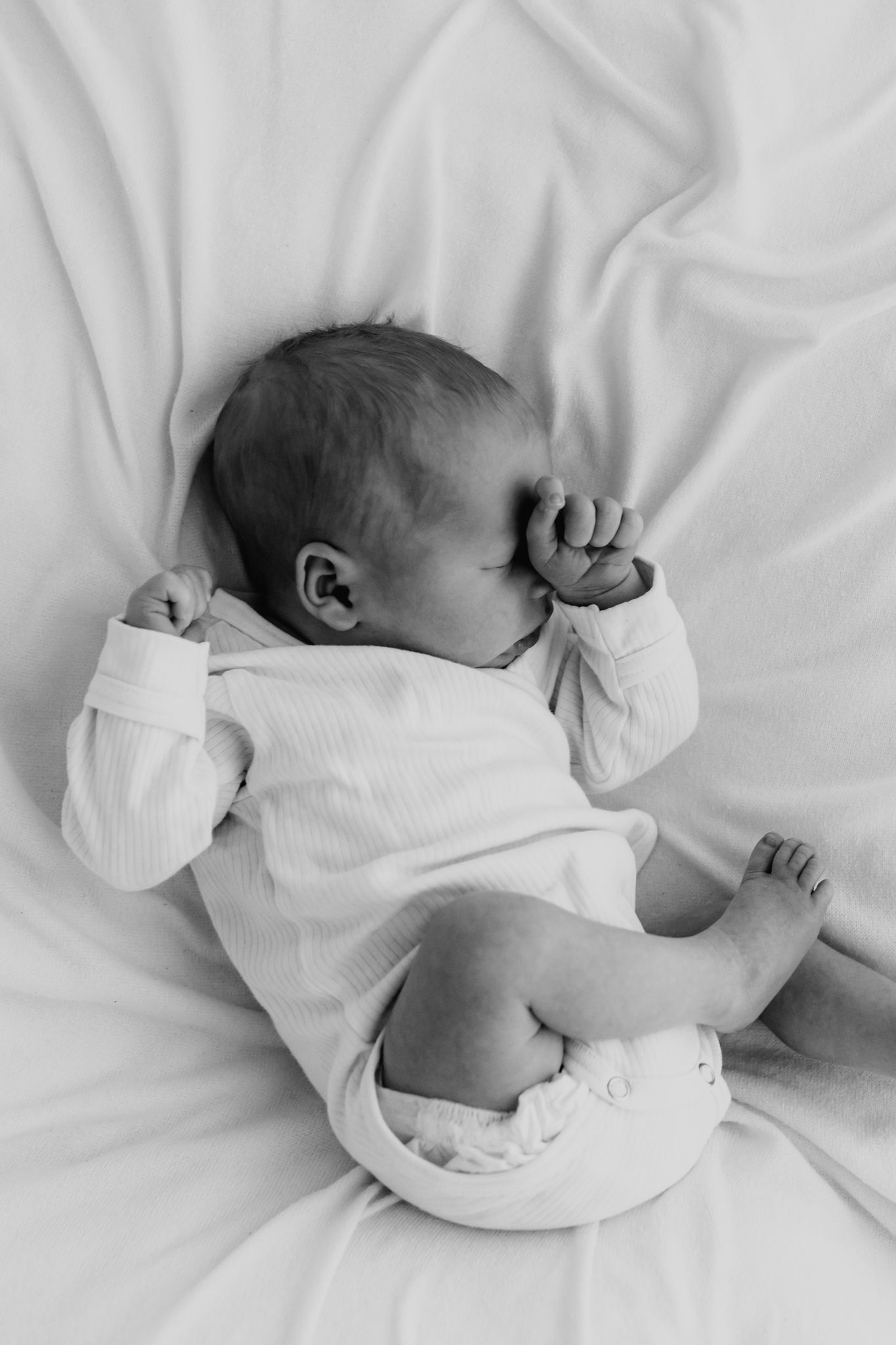 Black and white photo of a sleeping newborn baby in a white onesie on soft white bedding.