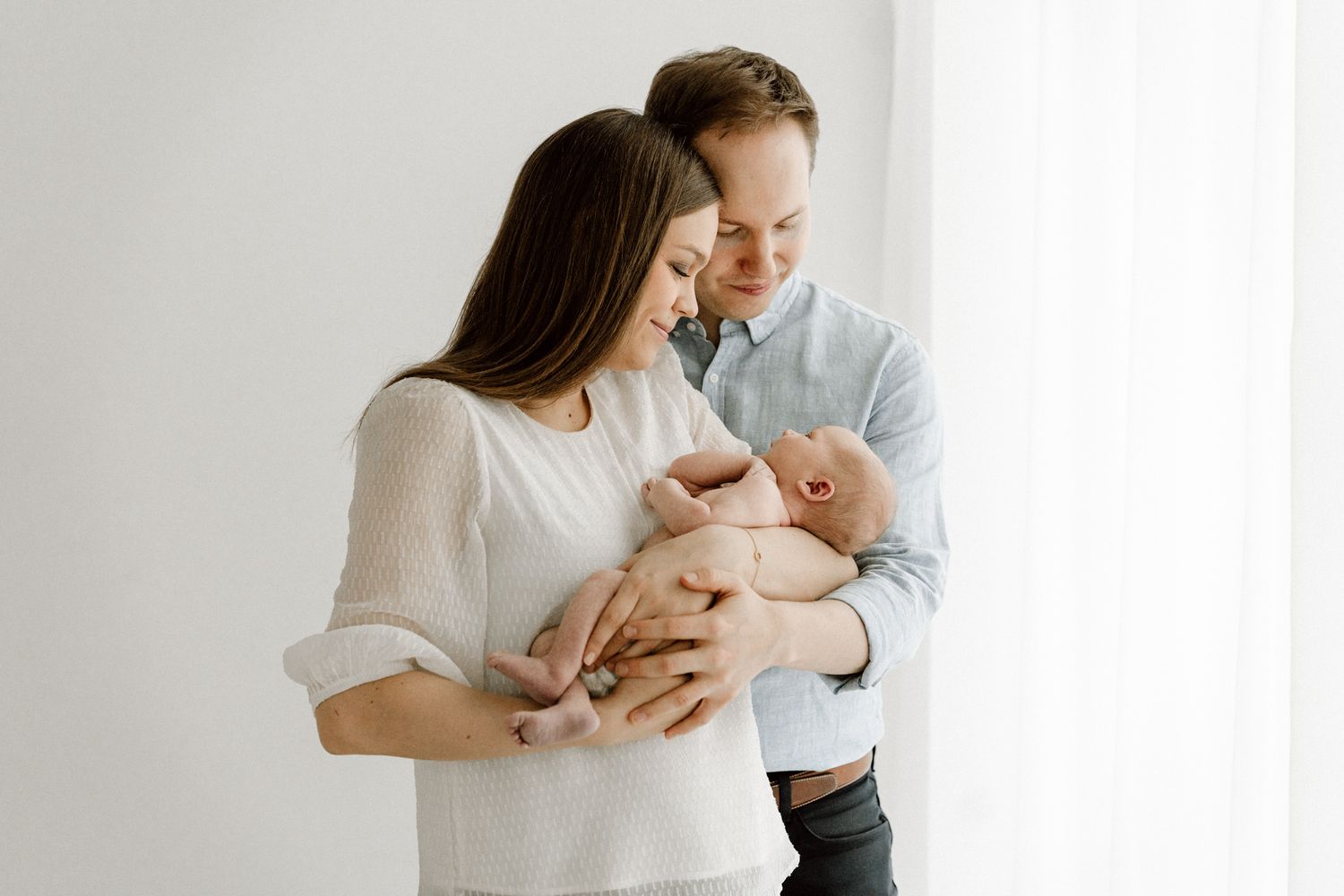Loving couple standing by bright window gently holding their newborn baby together.