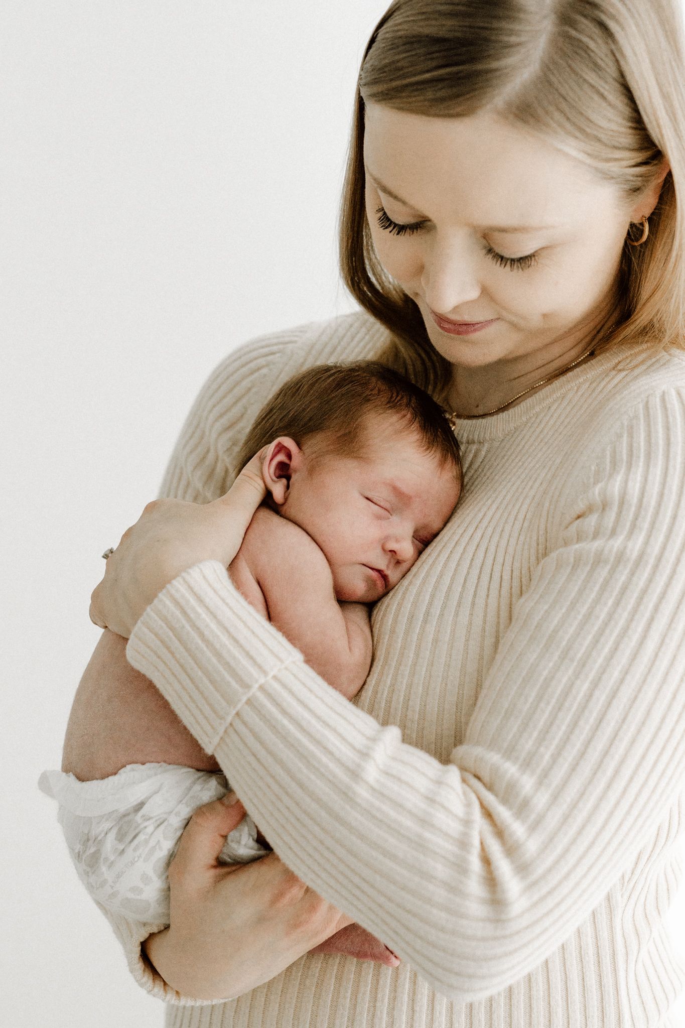 Smiling blonde mother in beige sweater cradles sleeping newborn baby close to chest.