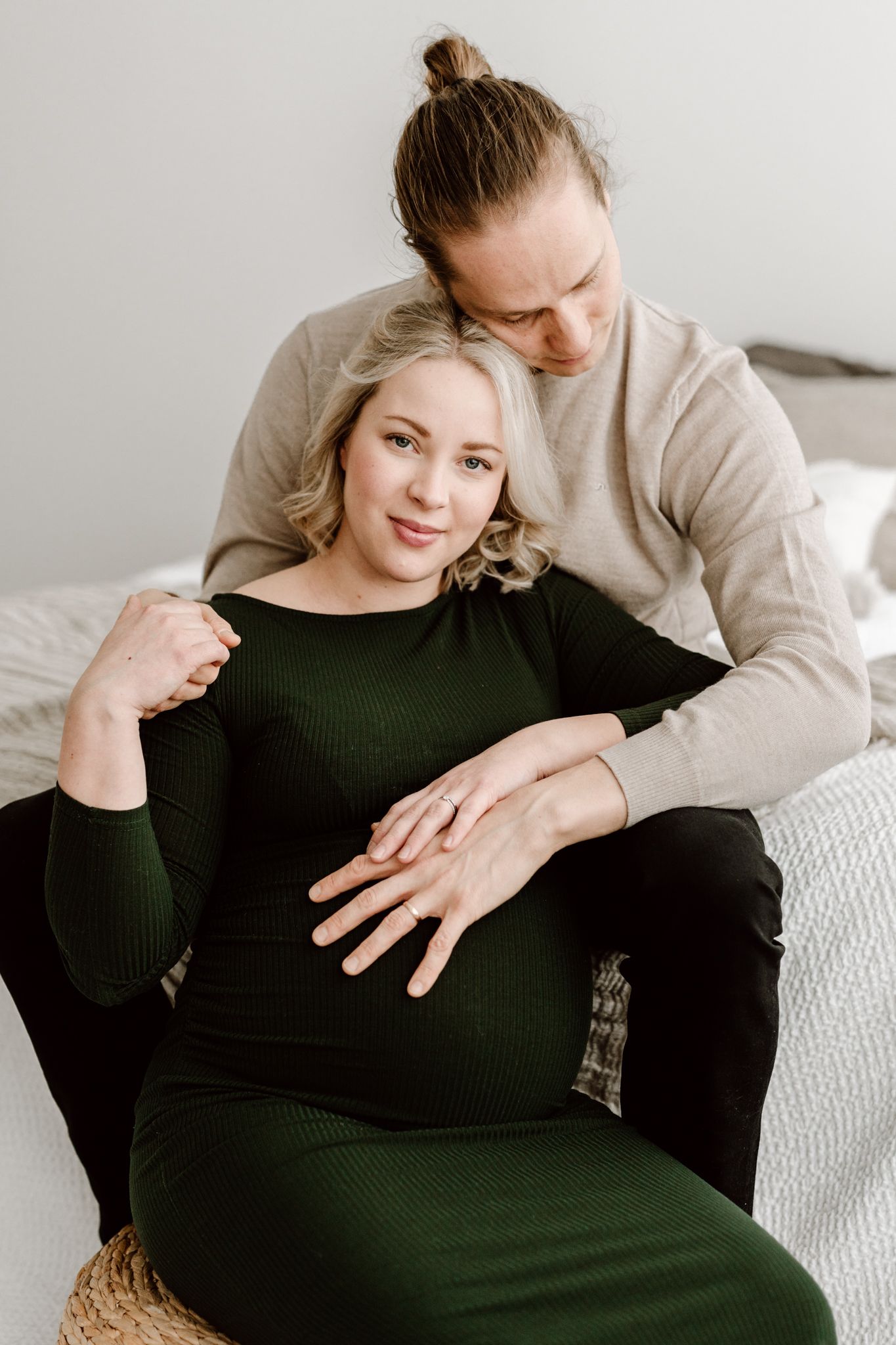 Pregnant woman in green dress leaning against partner while he cradles her baby bump on bed.