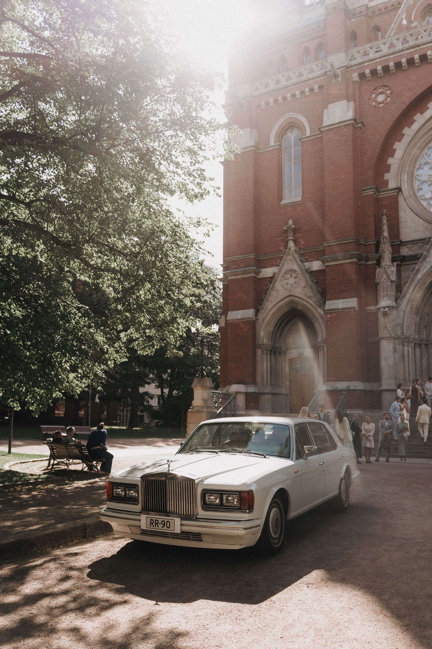 White Rolls-Royce wedding car parked outside a Gothic red brick church on a sunny day.