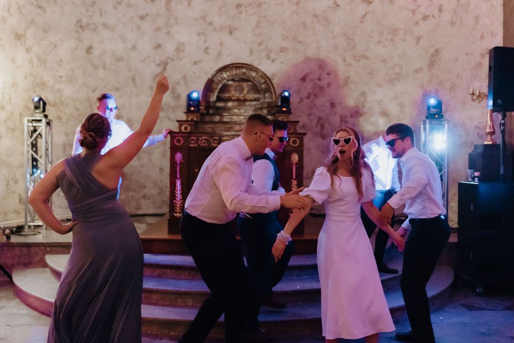 Wedding party performs energetic choreographed dance with bride and groom wearing sunglasses at reception.