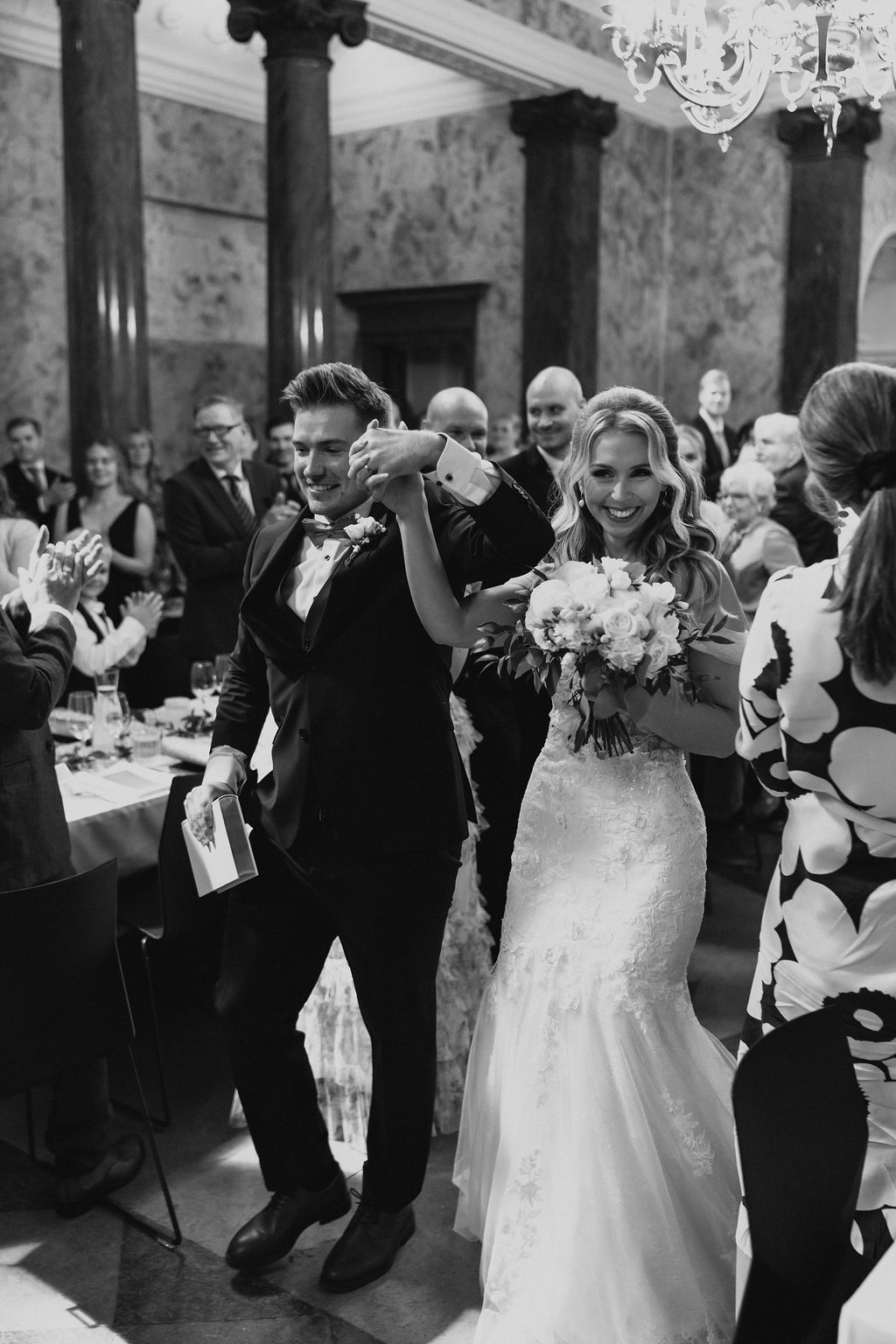Black and white photo of newlyweds celebrating recessional with raised hands among guests.