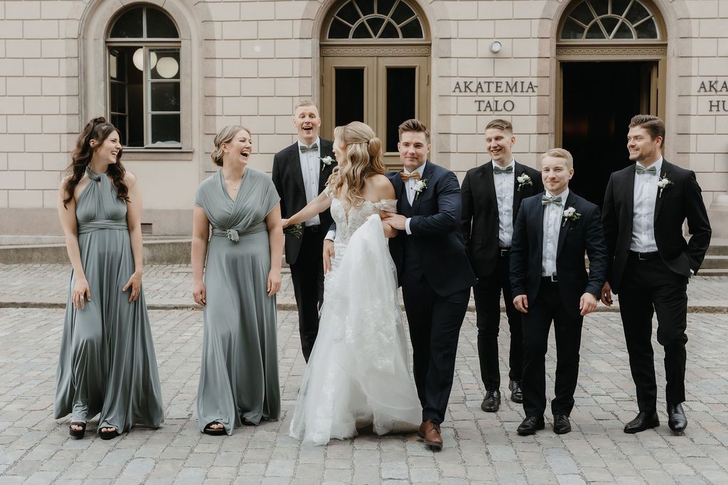 Laughing wedding party strolls past Akatemia Talo with bridesmaids in sage green gowns and groomsmen in black.