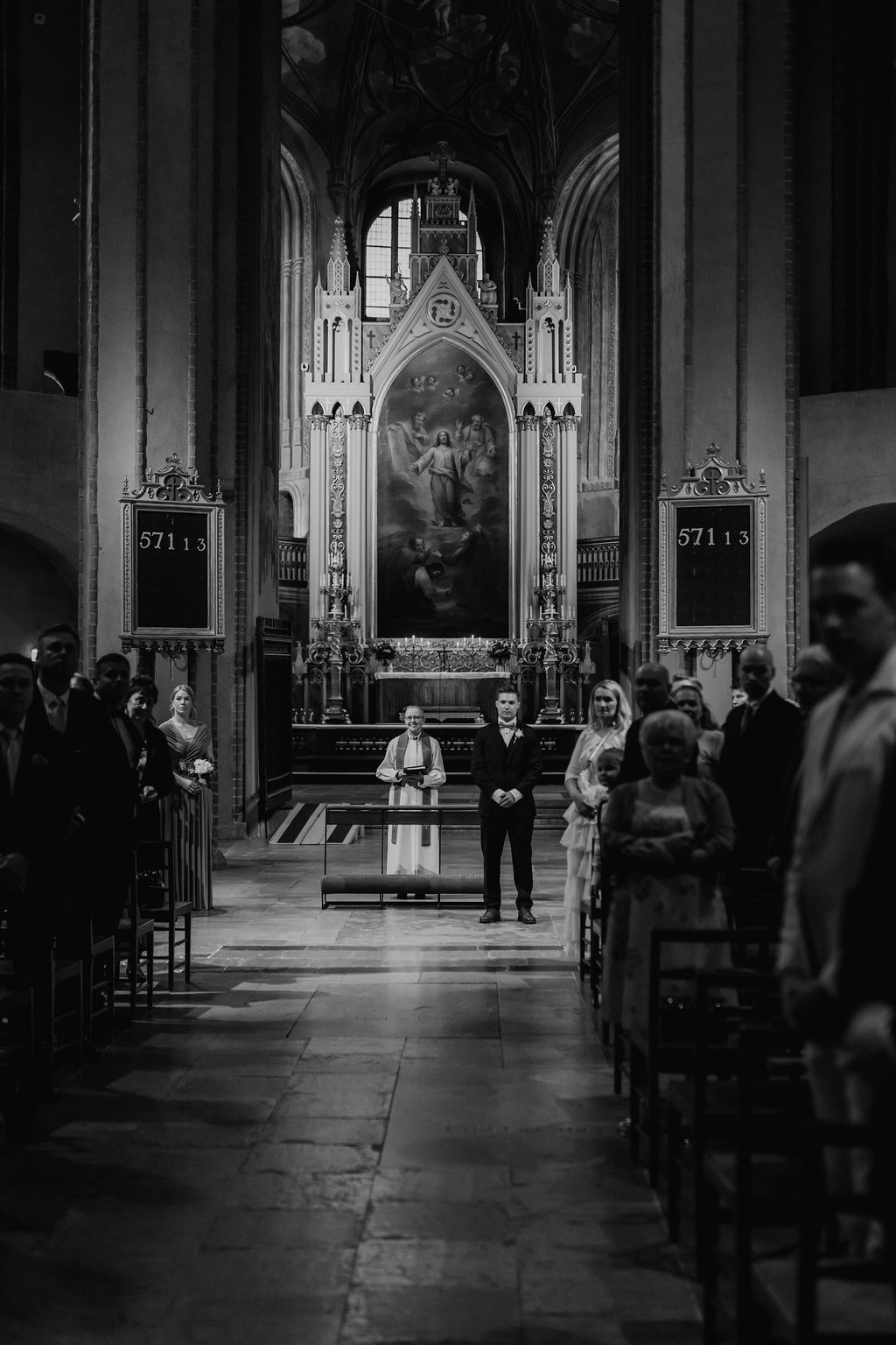 Bride and groom stand before ornate altar in a grand Gothic church in black and white.