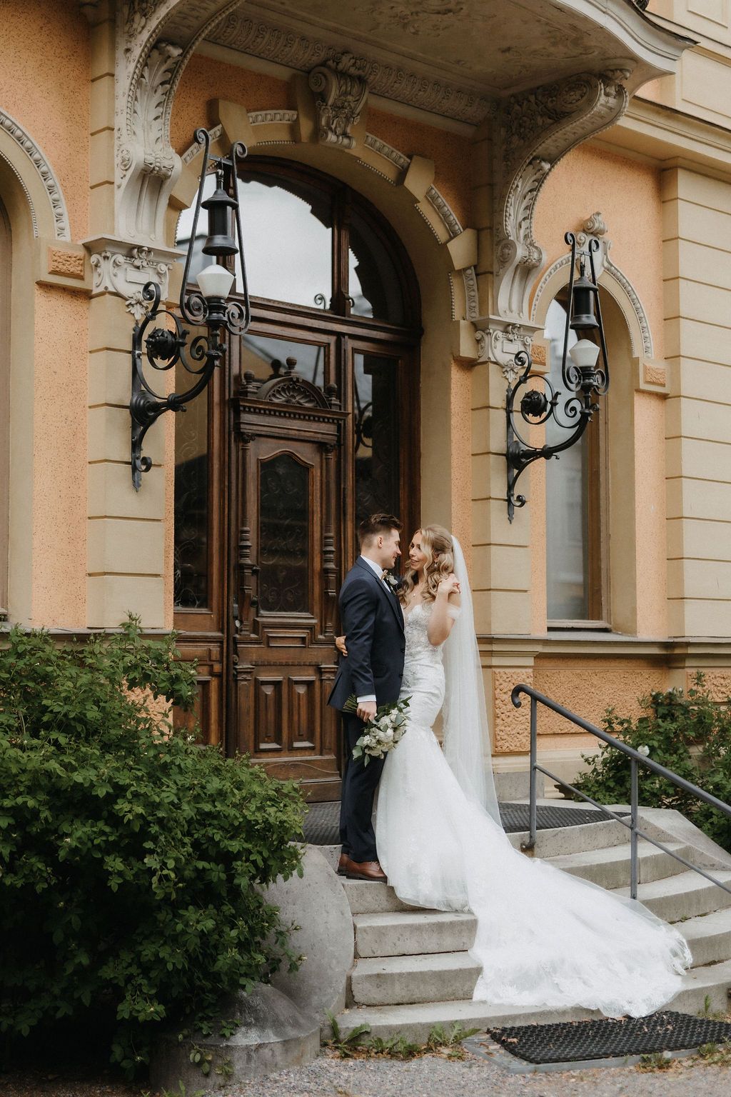 Bride and groom share romantic moment on steps of ornate Art Nouveau building with decorative iron lanterns.