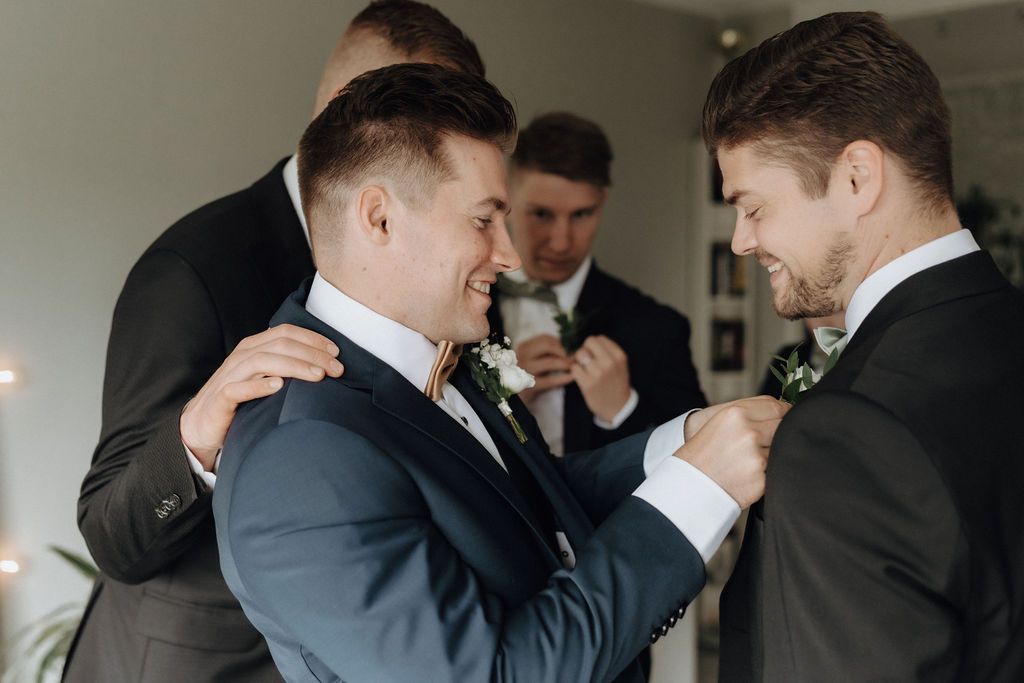 Groomsmen help each other with boutonnieres and suits before a wedding ceremony.
