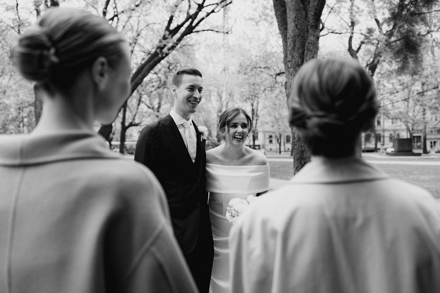 Bride and groom smiling at guests outdoors in a park setting, captured in black and white.