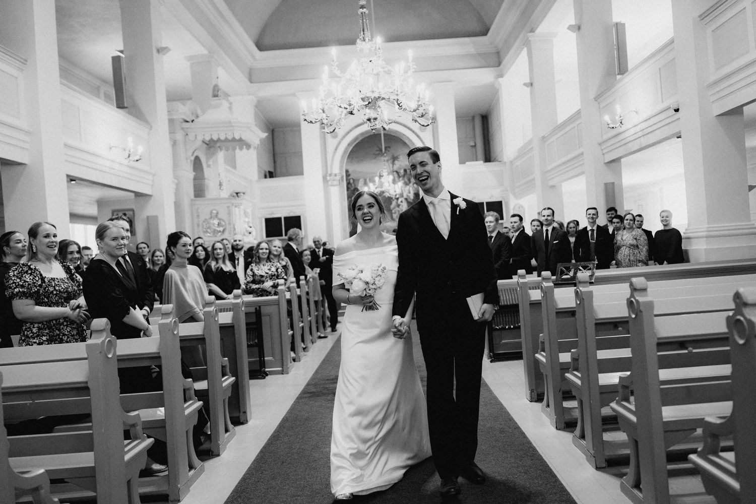 Bride and groom walk down the aisle of a grand chandelier-lit church after their wedding ceremony.