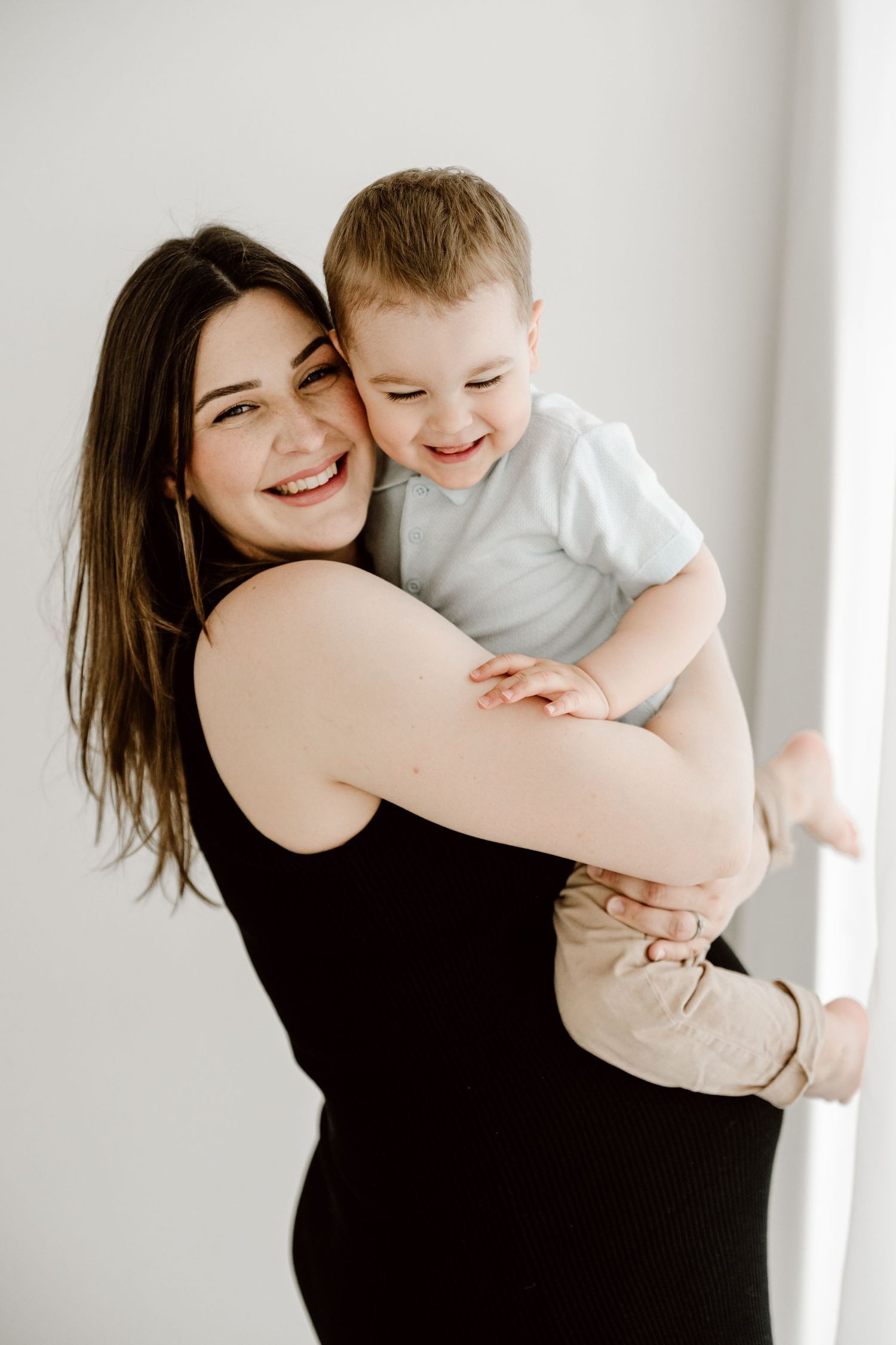 Smiling young mother holding laughing toddler boy in white onesie against light background.