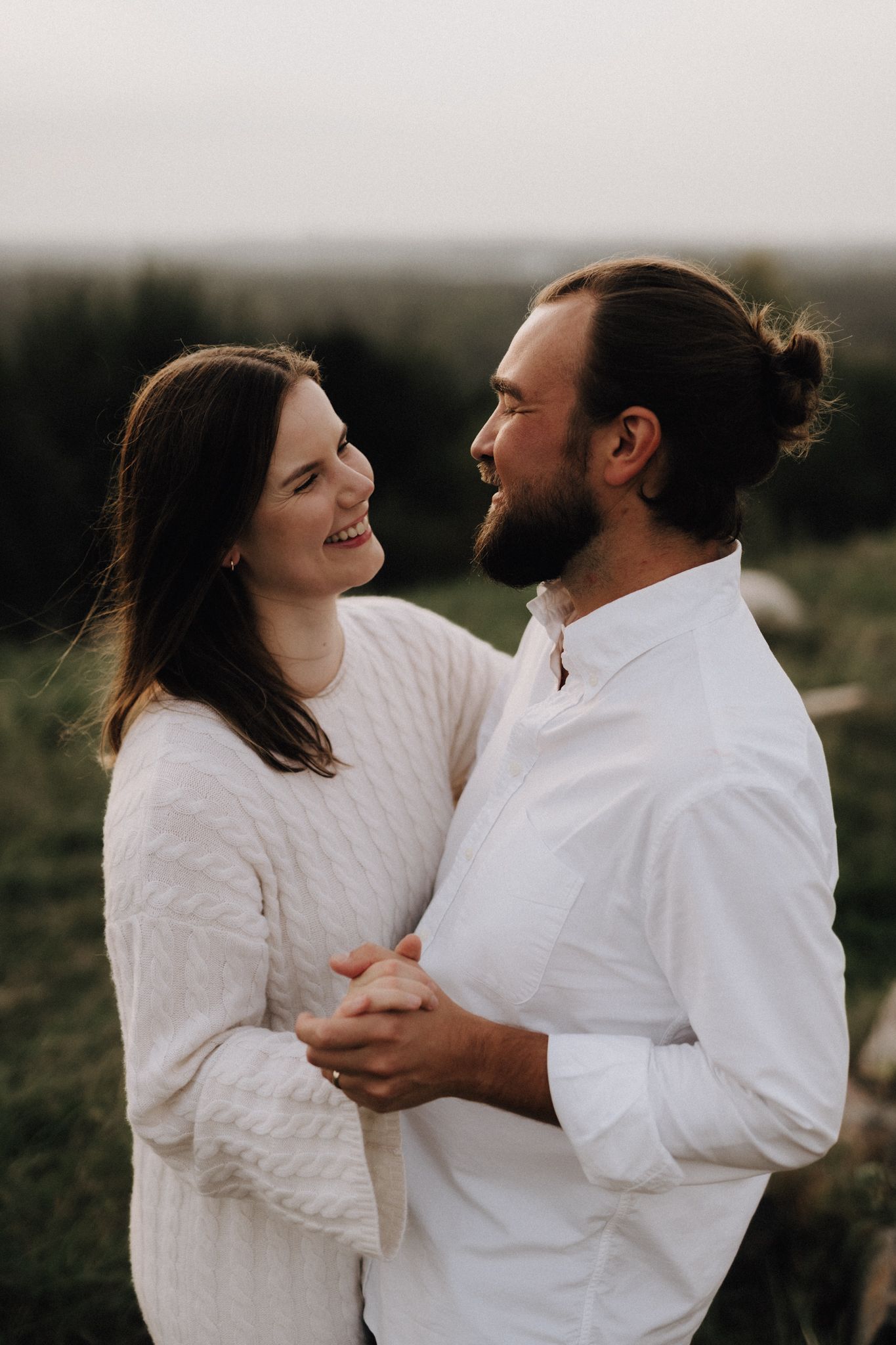 A smiling couple in white outfits embraces and gazes at each other lovingly in a natural outdoor setting.