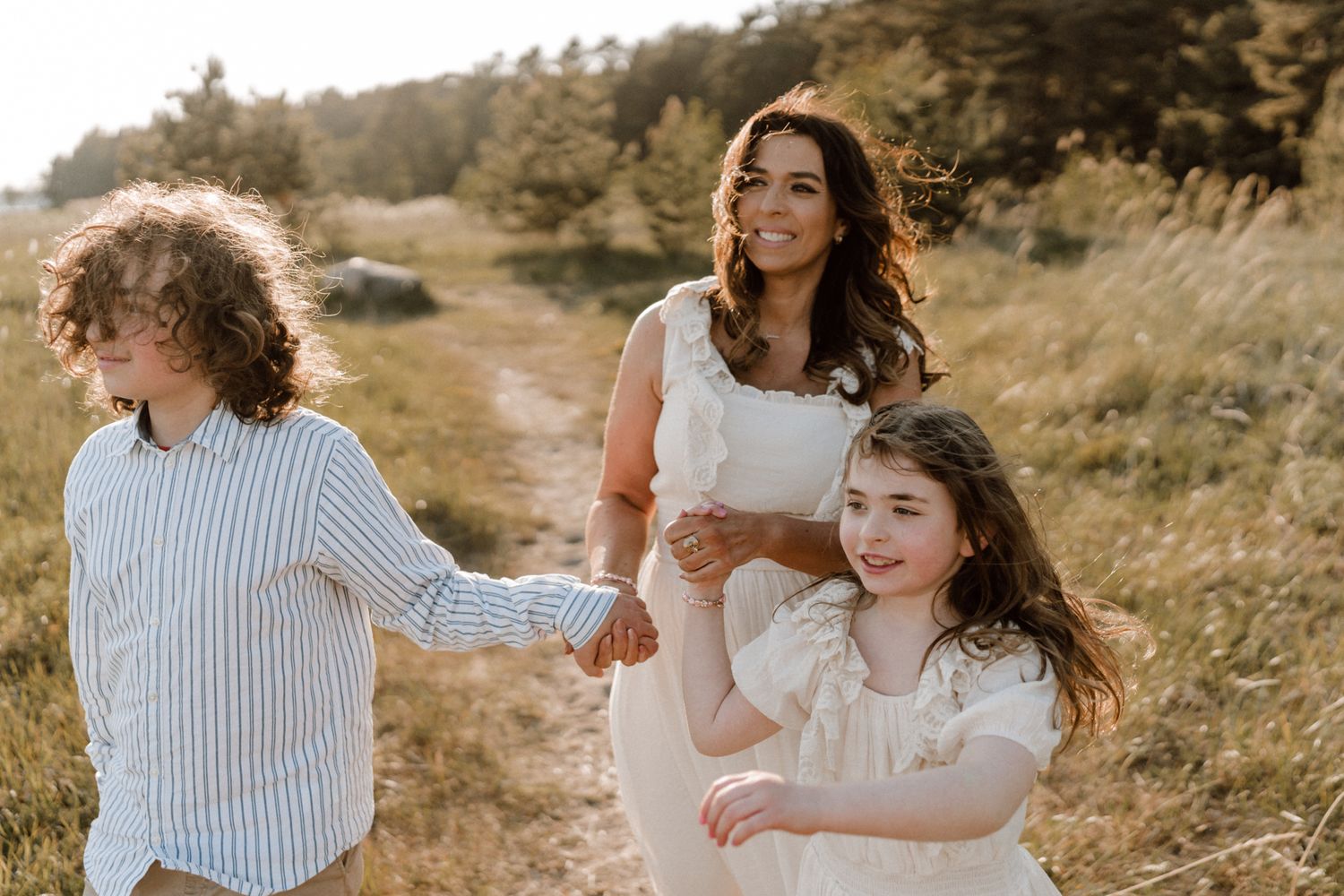 A smiling mother in a white dress walks hand-in-hand with her two children through a sunny meadow.