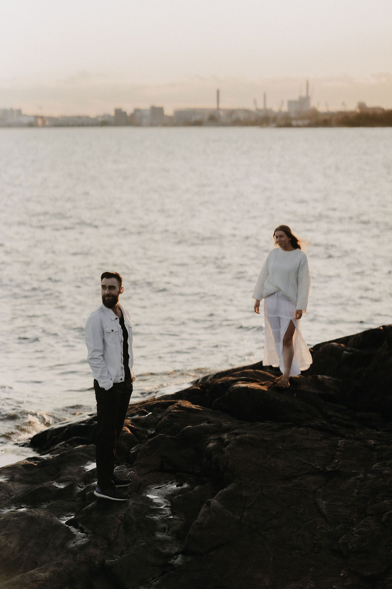 Bearded man in white jacket and woman in white dress posing on rocks by the sea at dusk.