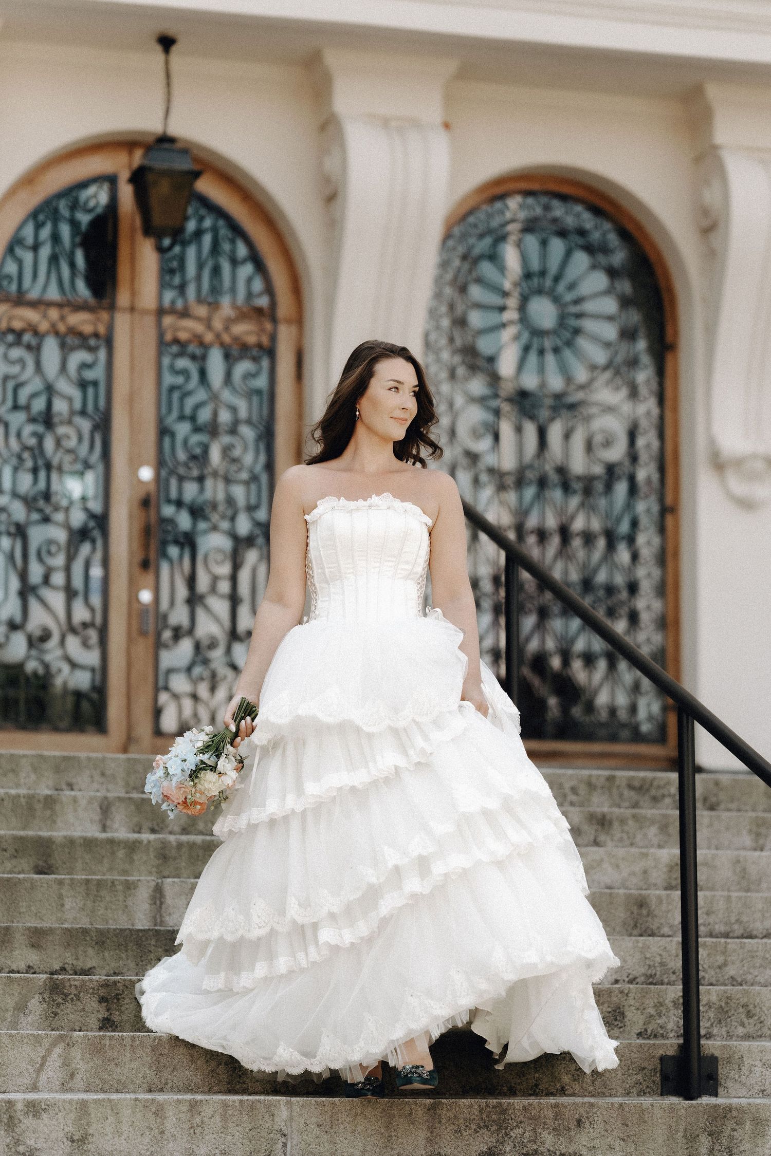 Elegant bride in a strapless tiered white ballgown holds a bouquet on stone steps before ornate doors.