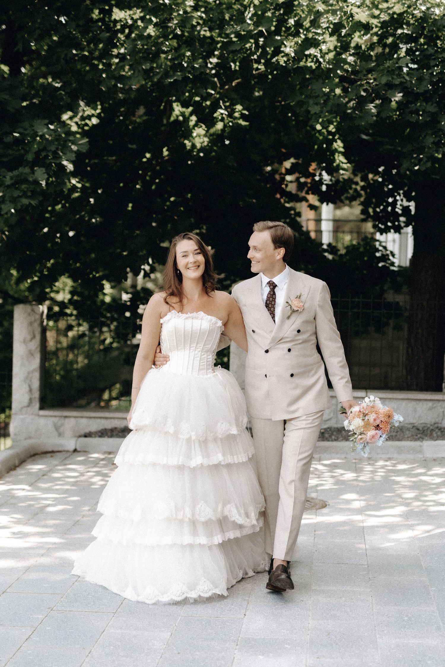 Smiling bride in white ruffled strapless gown walks with groom in beige suit holding bouquet outdoors.