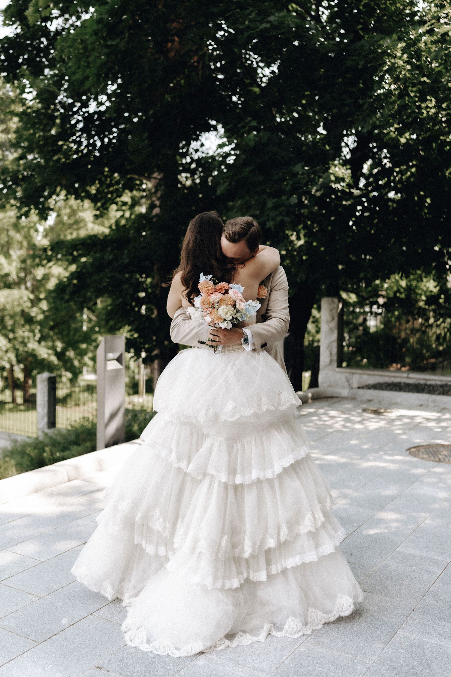 Wedding couple sharing a tender kiss, bride in layered white ruffled gown holding peach and blue bouquet in park.