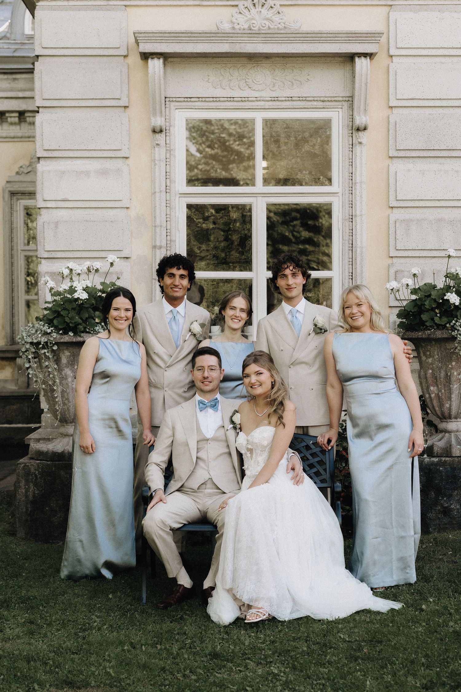 Bride and groom seated with smiling wedding party in coordinated blue-grey and beige outfits outside elegant venue.