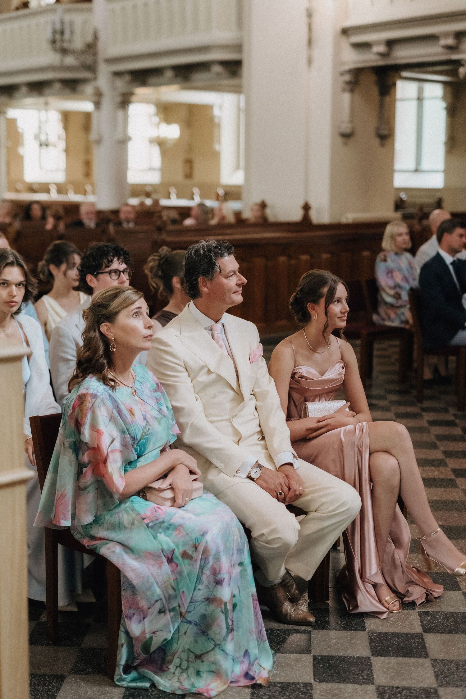 Wedding guests in formal attire seated in church pews during ceremony, woman in floral dress prominent.