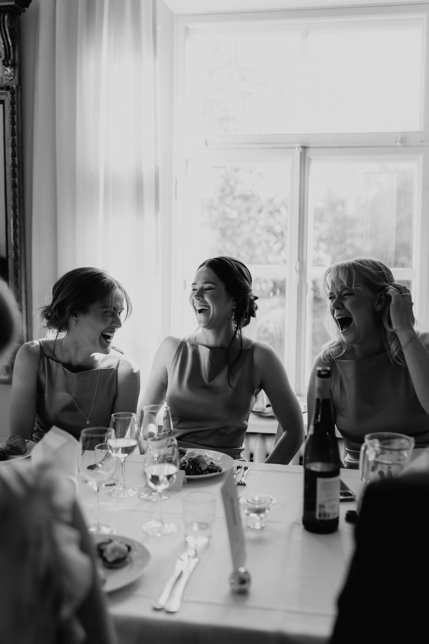 Black and white photo of three bridesmaids laughing joyfully together at a wedding reception dinner table.