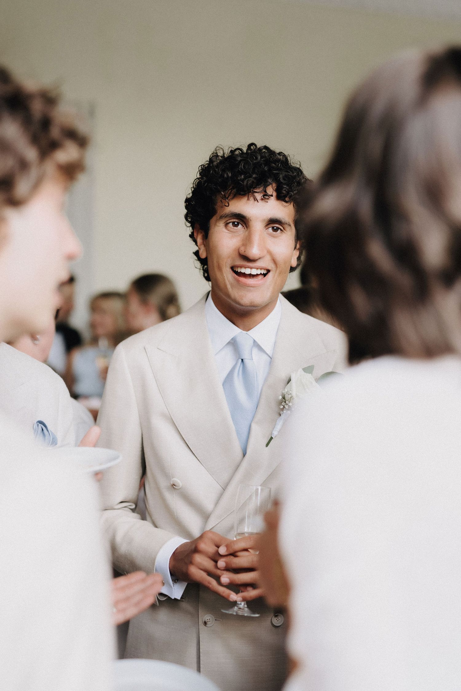 Smiling groom in beige double-breasted suit with boutonniere holds champagne glass at wedding reception.