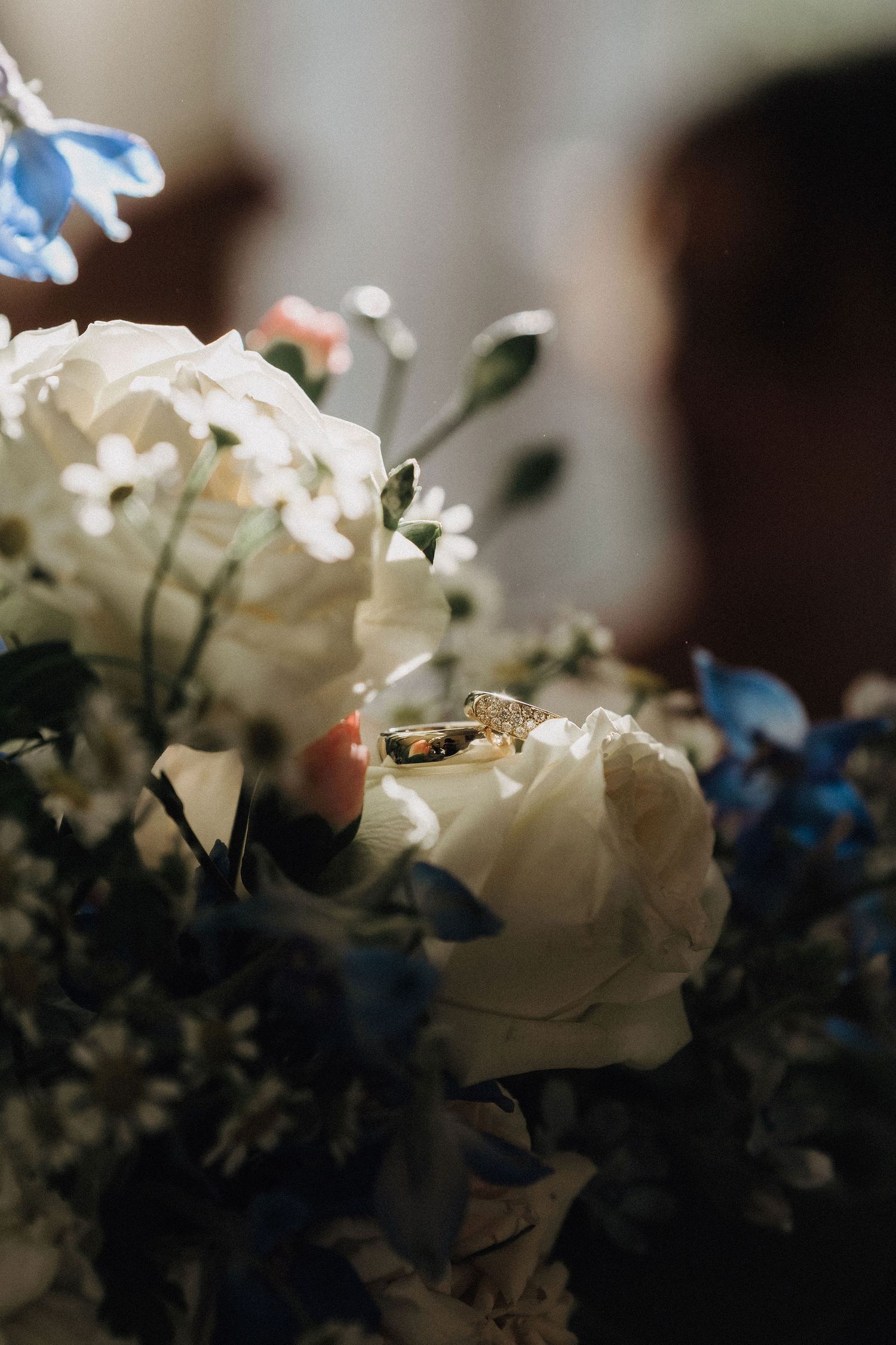 Gold diamond wedding ring resting on white roses in a bridal bouquet with blue flowers.