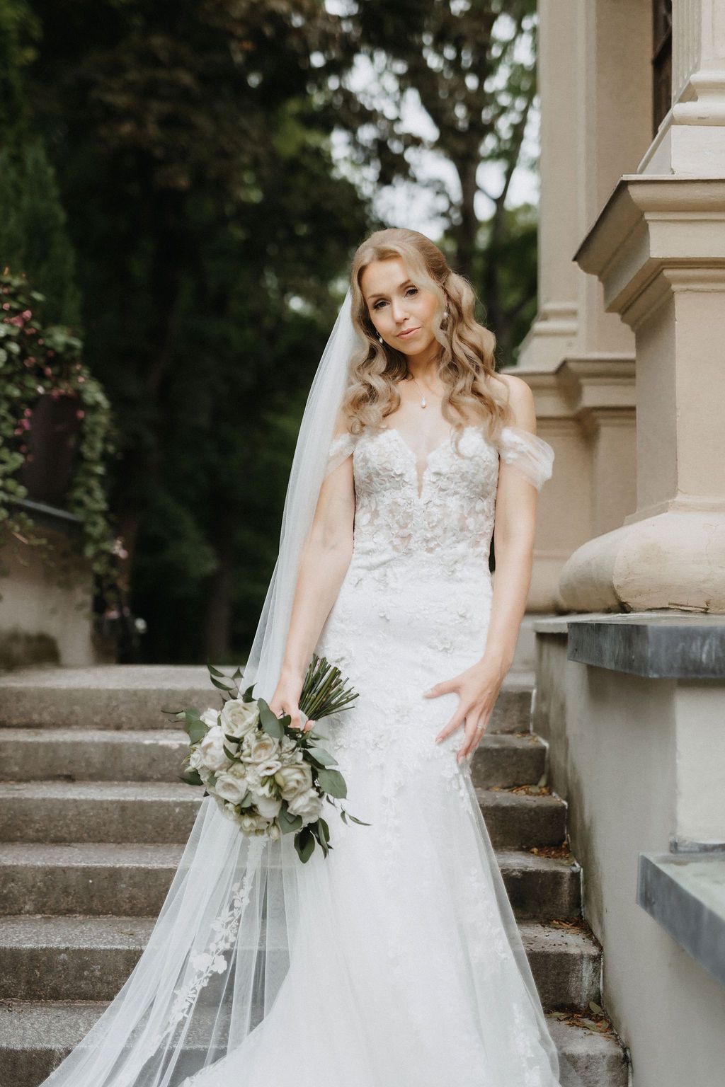 Blonde bride in off-shoulder lace mermaid dress with cathedral veil poses on stone staircase.