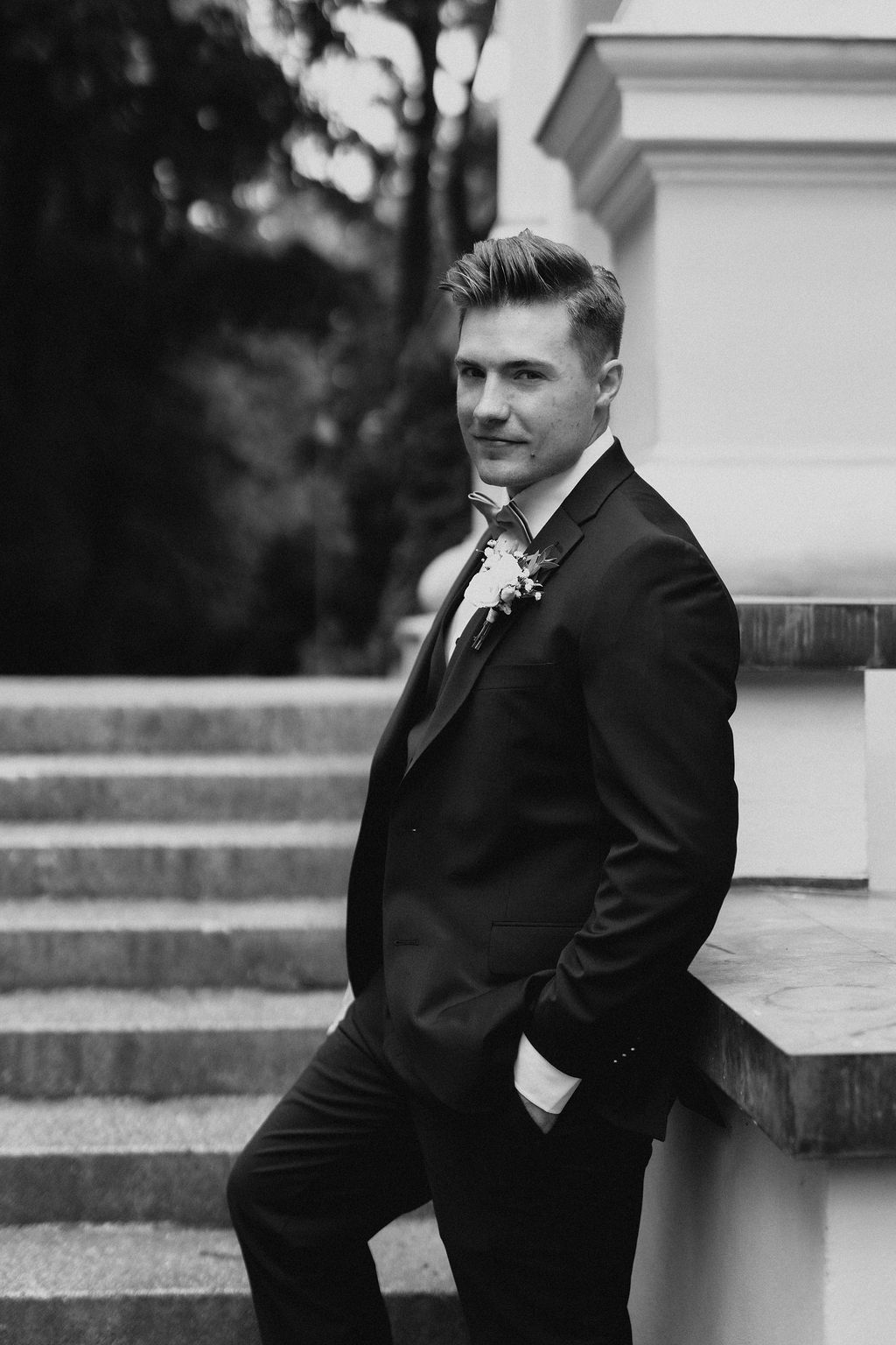 Elegant groom in black tuxedo leaning against stone wall near staircase, black and white wedding portrait.