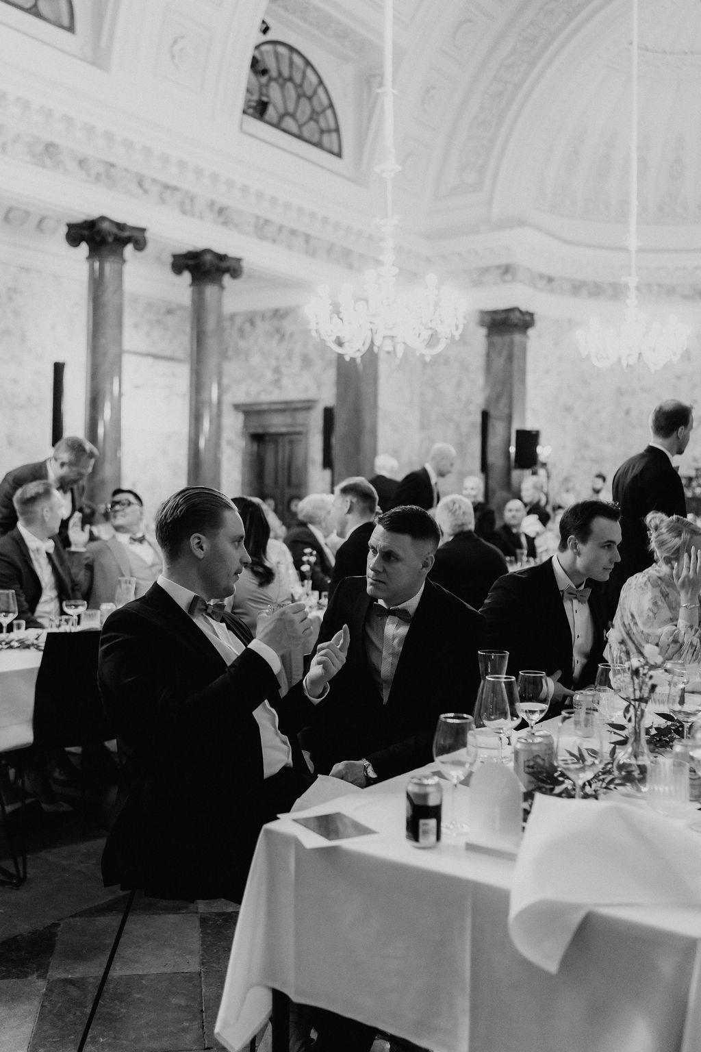 Wedding guests in tuxedos seated at banquet table in elegant neoclassical ballroom with chandelier.