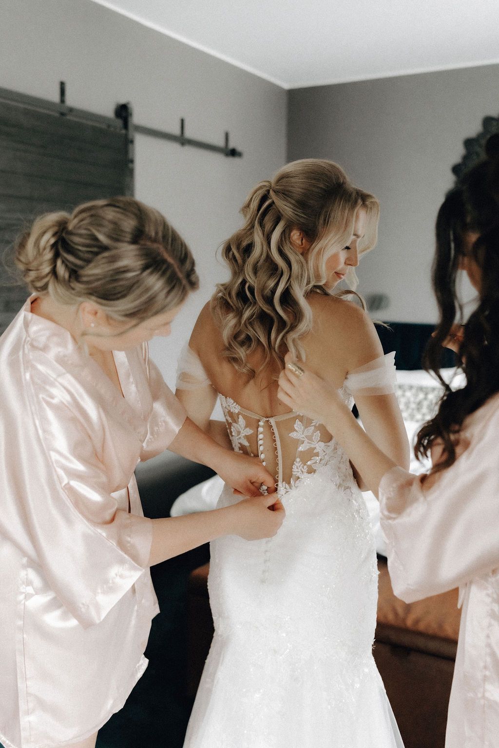 Bridesmaids in pink robes helping bride button up backless lace wedding gown.
