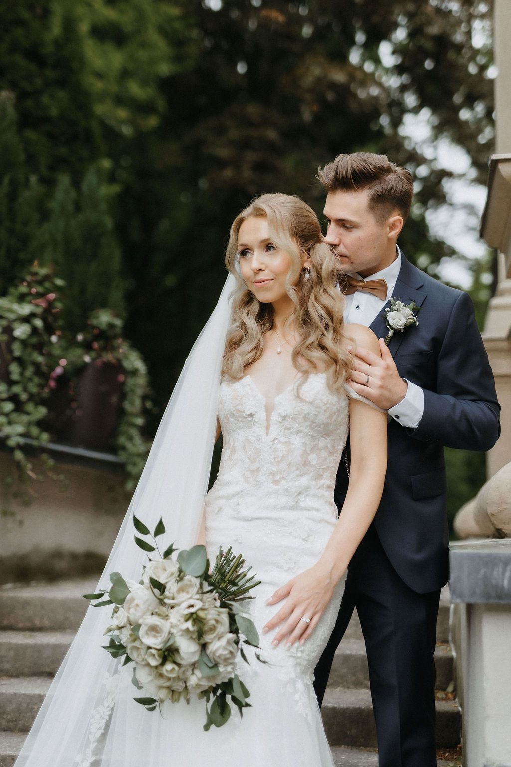 Groom in navy suit embraces bride in lace mermaid gown while kissing her cheek on stairs.