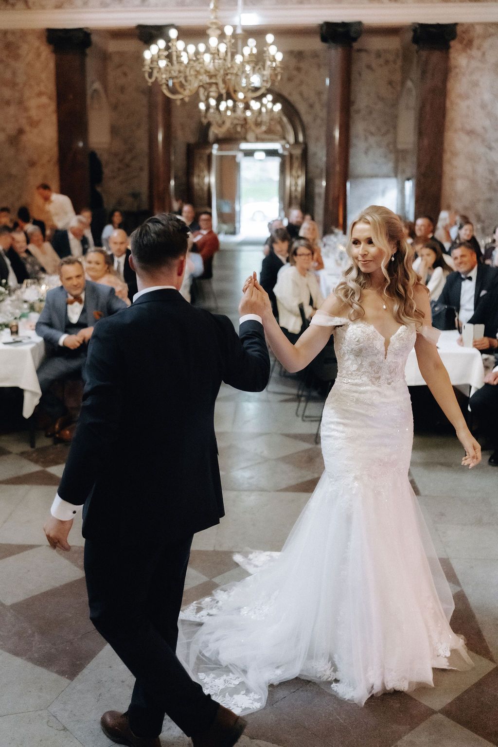 Bride and groom sharing first dance in grand marble ballroom with seated wedding guests.