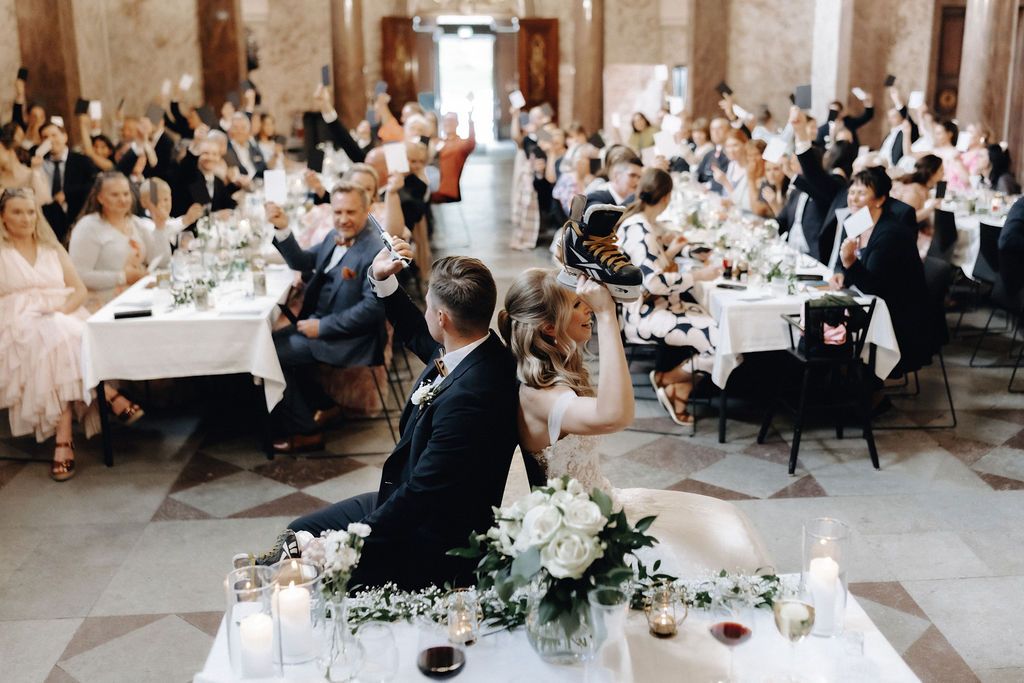 Bride and groom sit at sweetheart table adorned with white florals as guests celebrate.