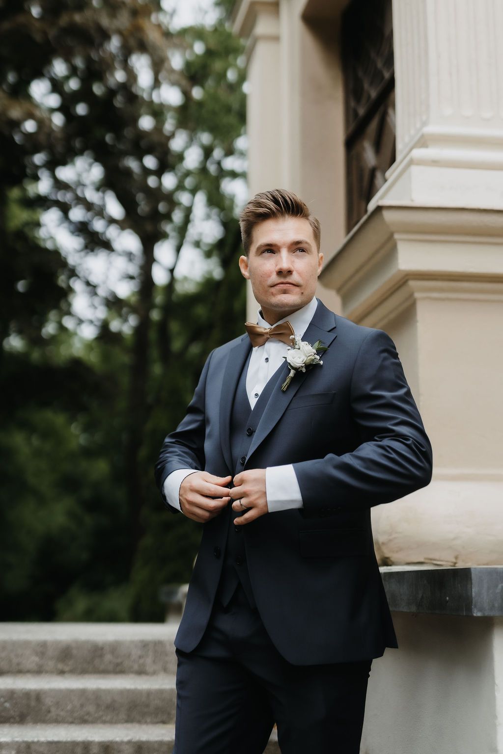 Dapper groom in navy three-piece suit with brown bow tie and boutonniere at outdoor wedding venue.
