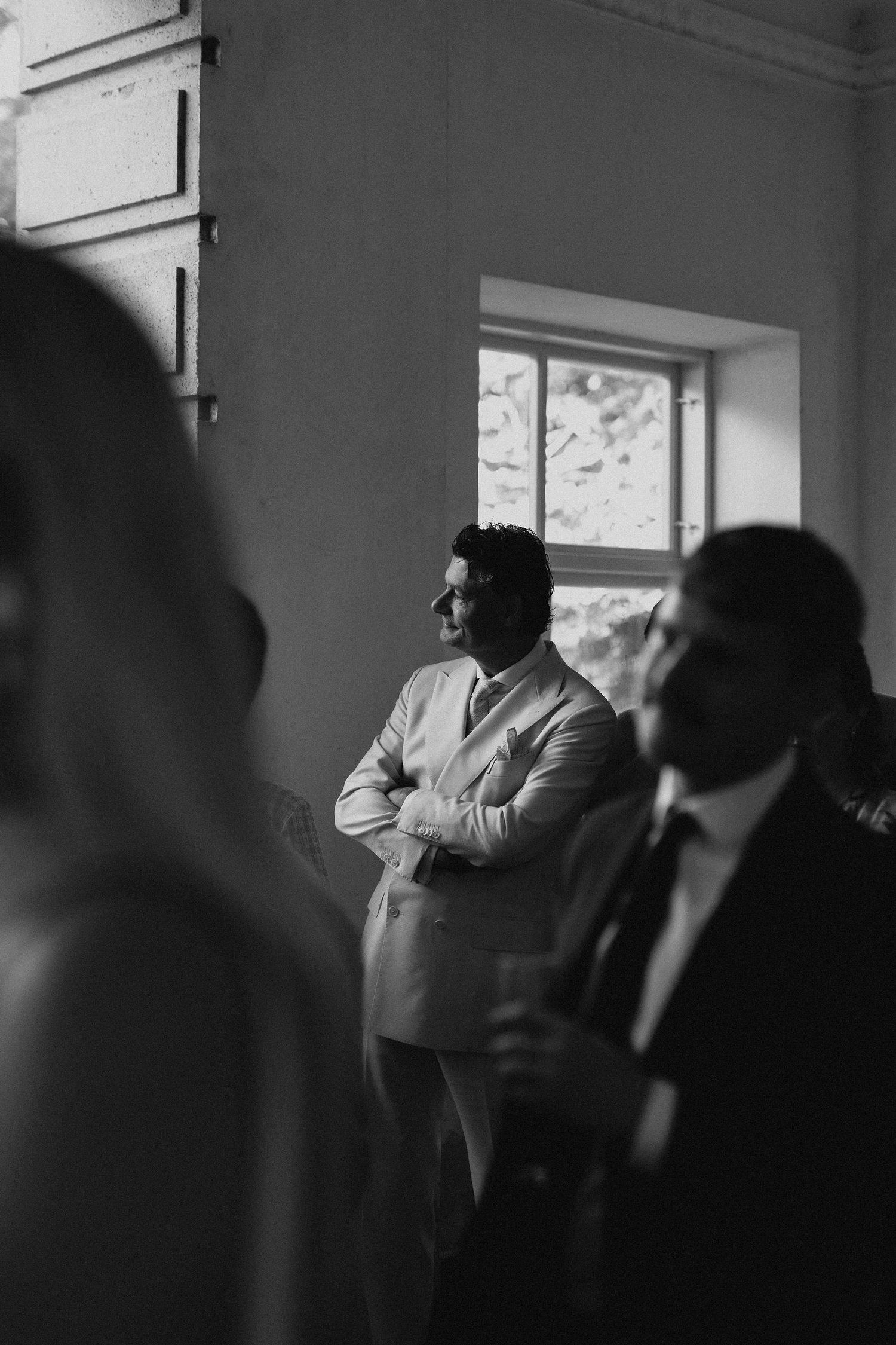 Black and white photo of groom in beige suit standing with arms crossed at wedding reception.