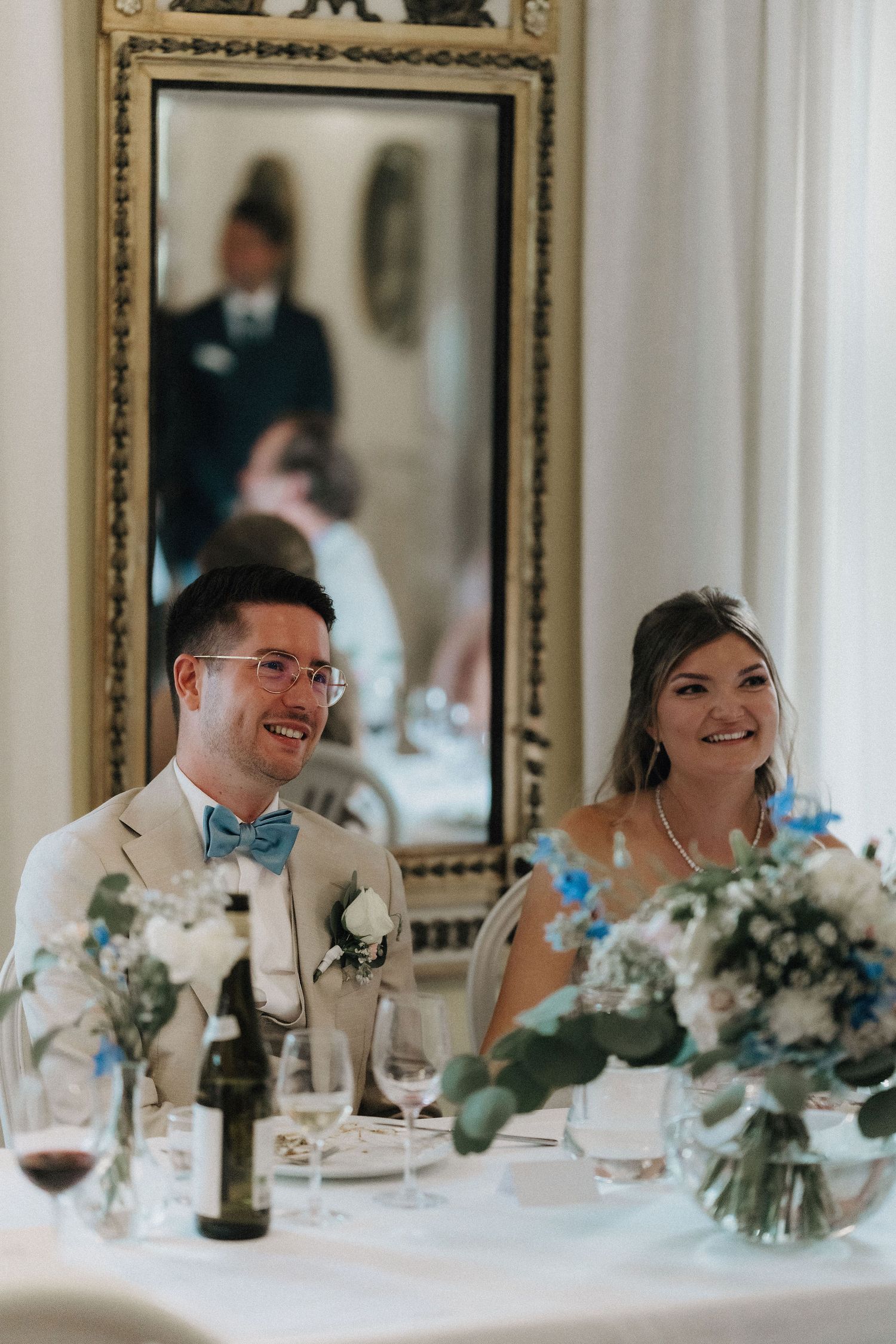 Smiling bride and groom in blue bow tie seated at head table during wedding reception.