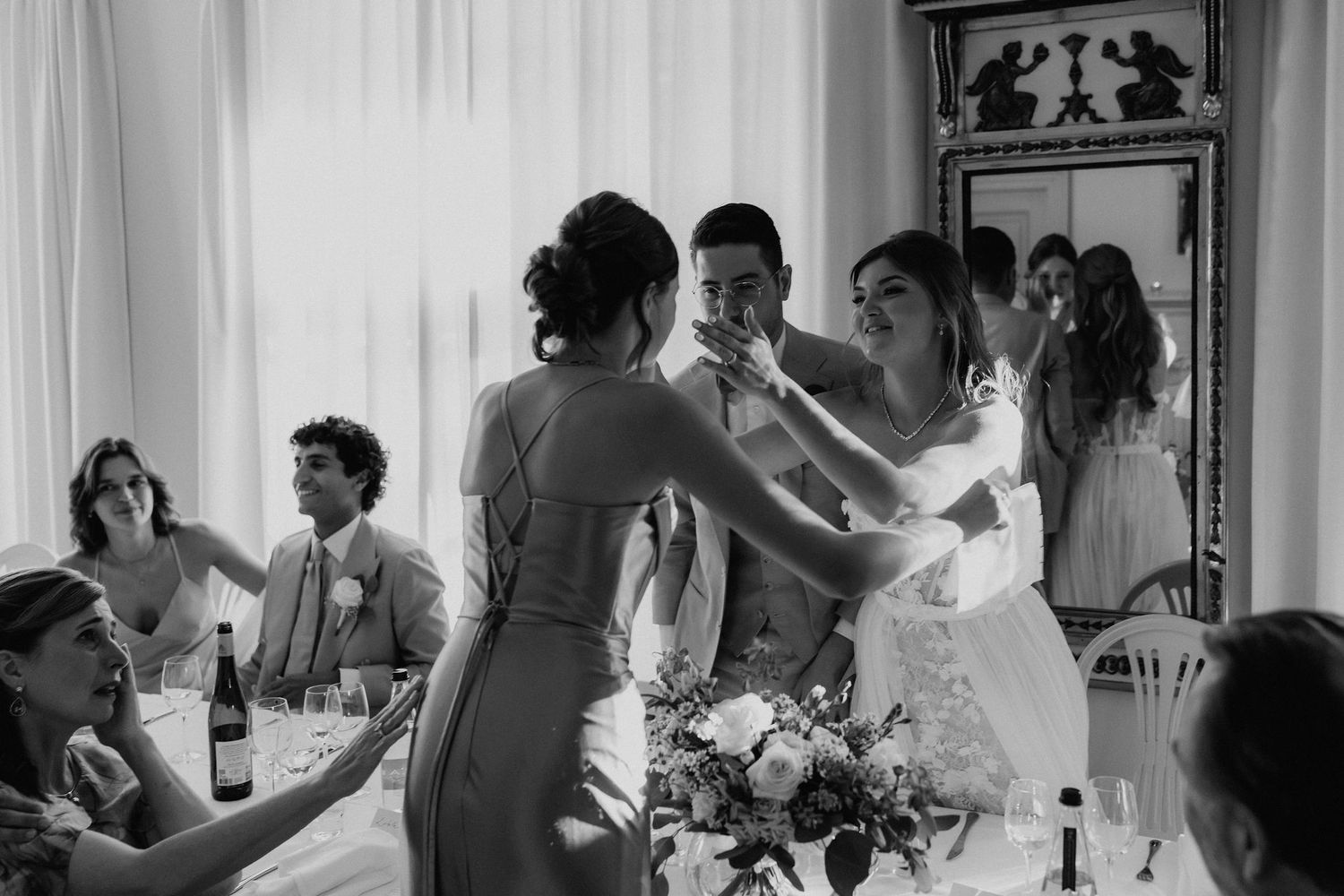 Black and white photo of bride and bridesmaid embracing at wedding reception dinner table.