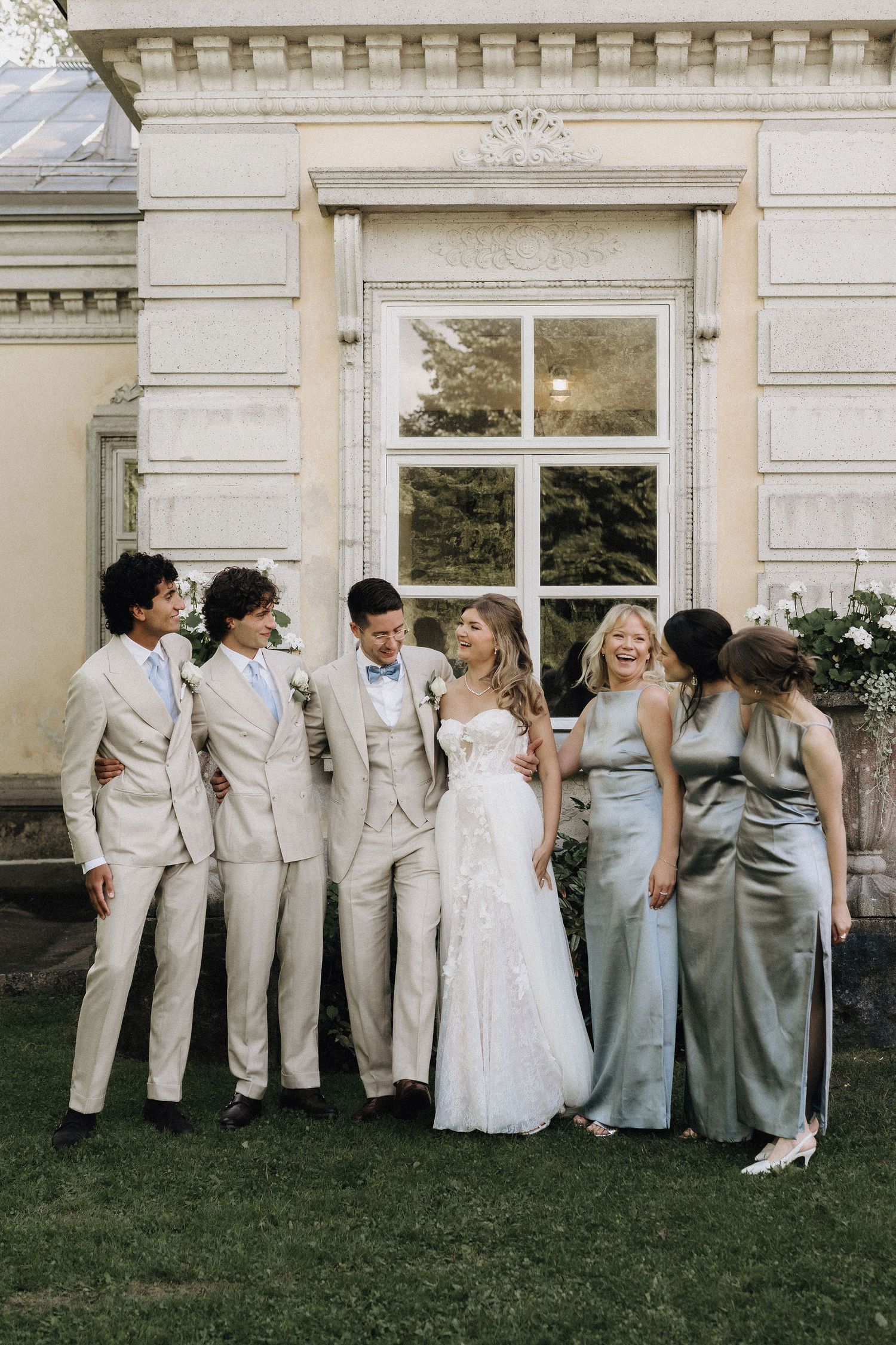 Bride and groom with wedding party sharing joyful moment on lawn outside ornate building with large window.