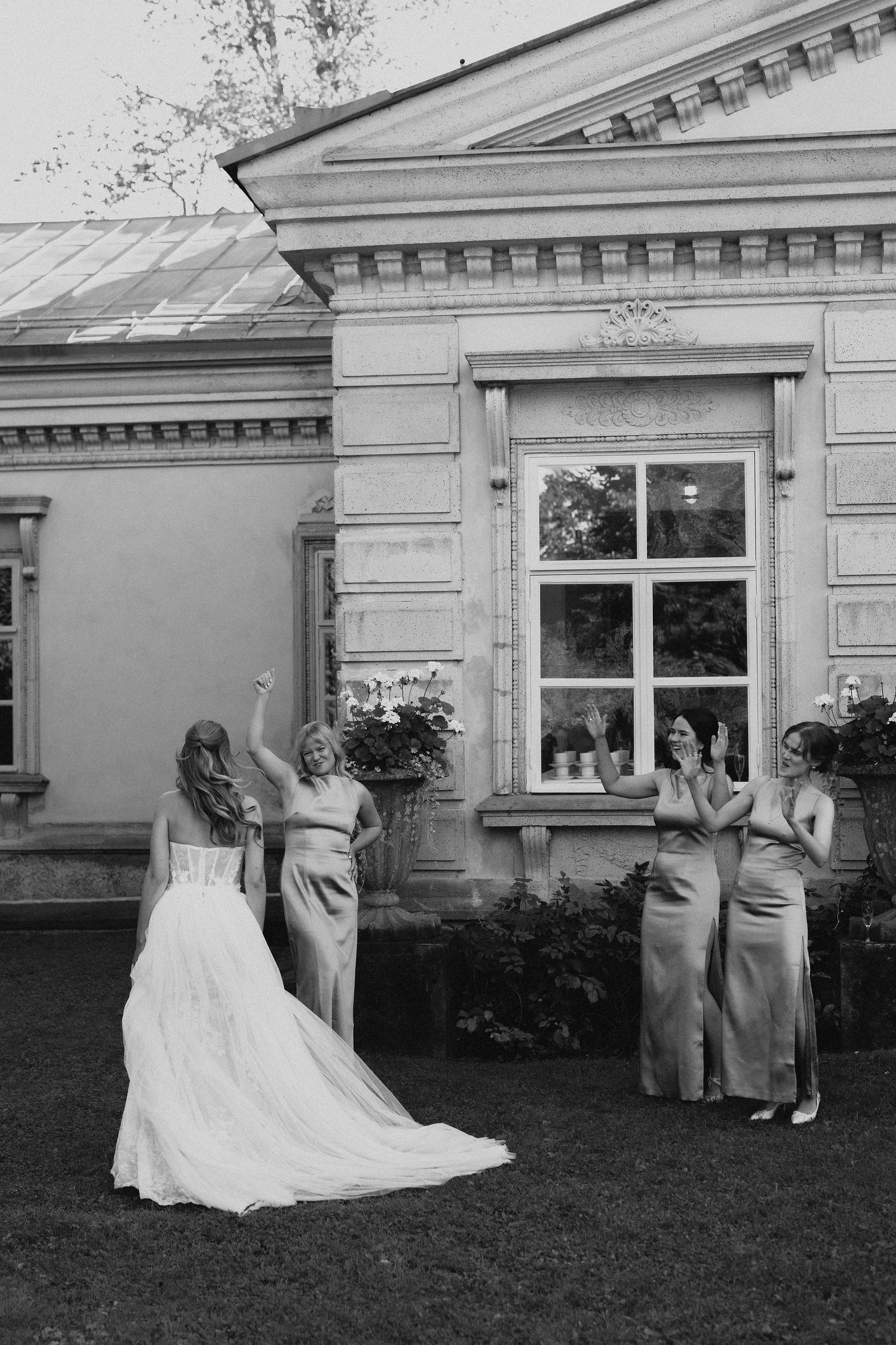 Bride raising fist triumphantly while bridesmaids in satin gowns cheer outside a classical building, black and white.