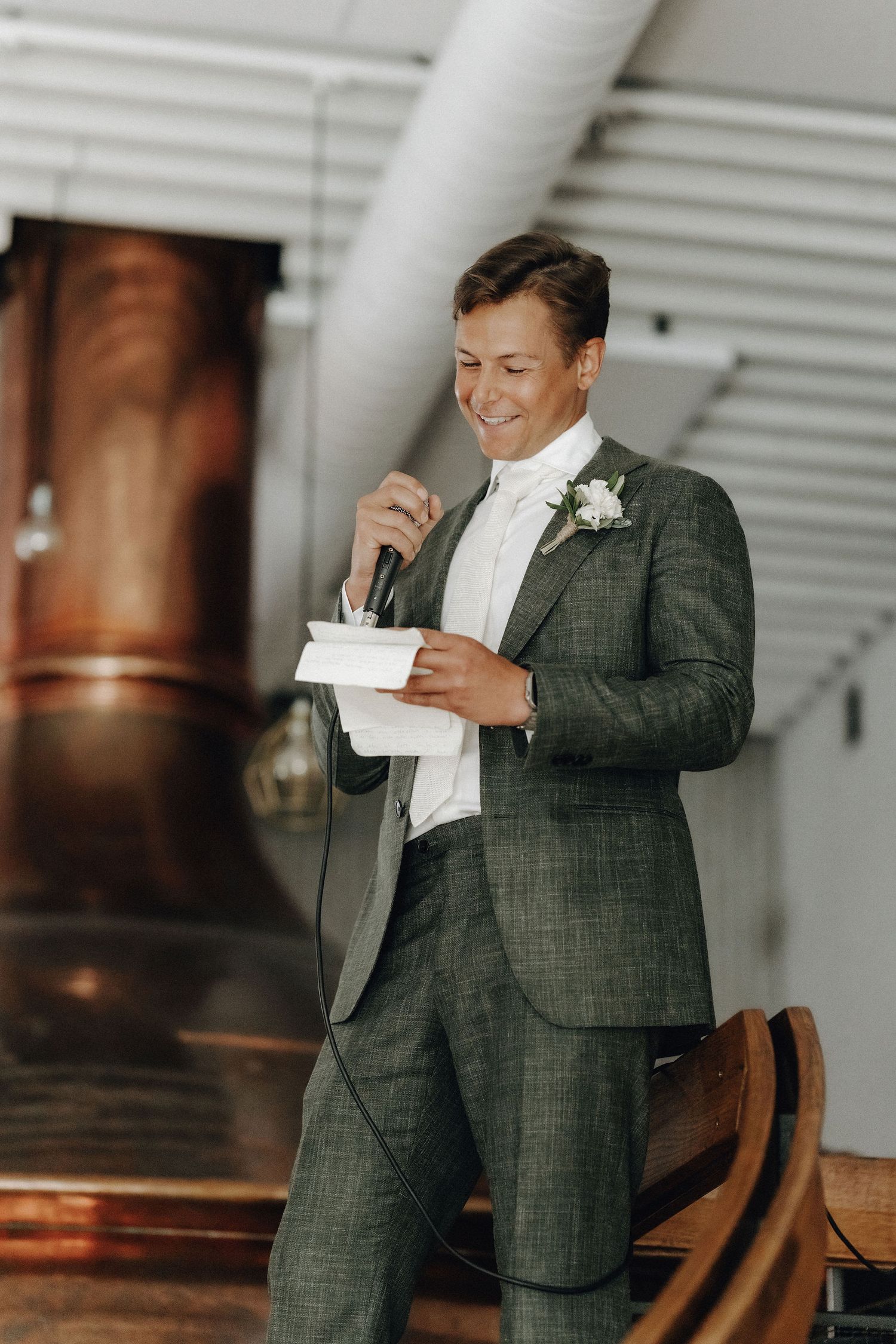 Smiling groom in green plaid suit delivering speech with microphone near copper brewery tanks.