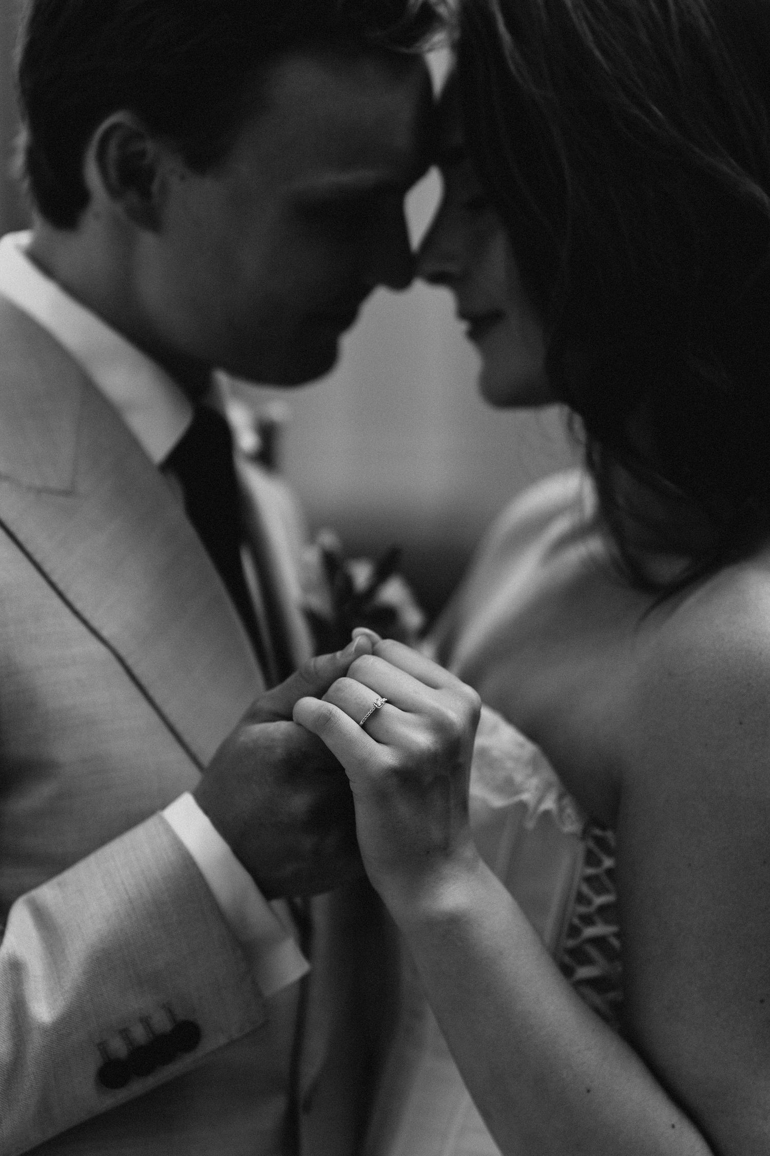 Black and white close-up of bride and groom touching foreheads while showcasing wedding ring.