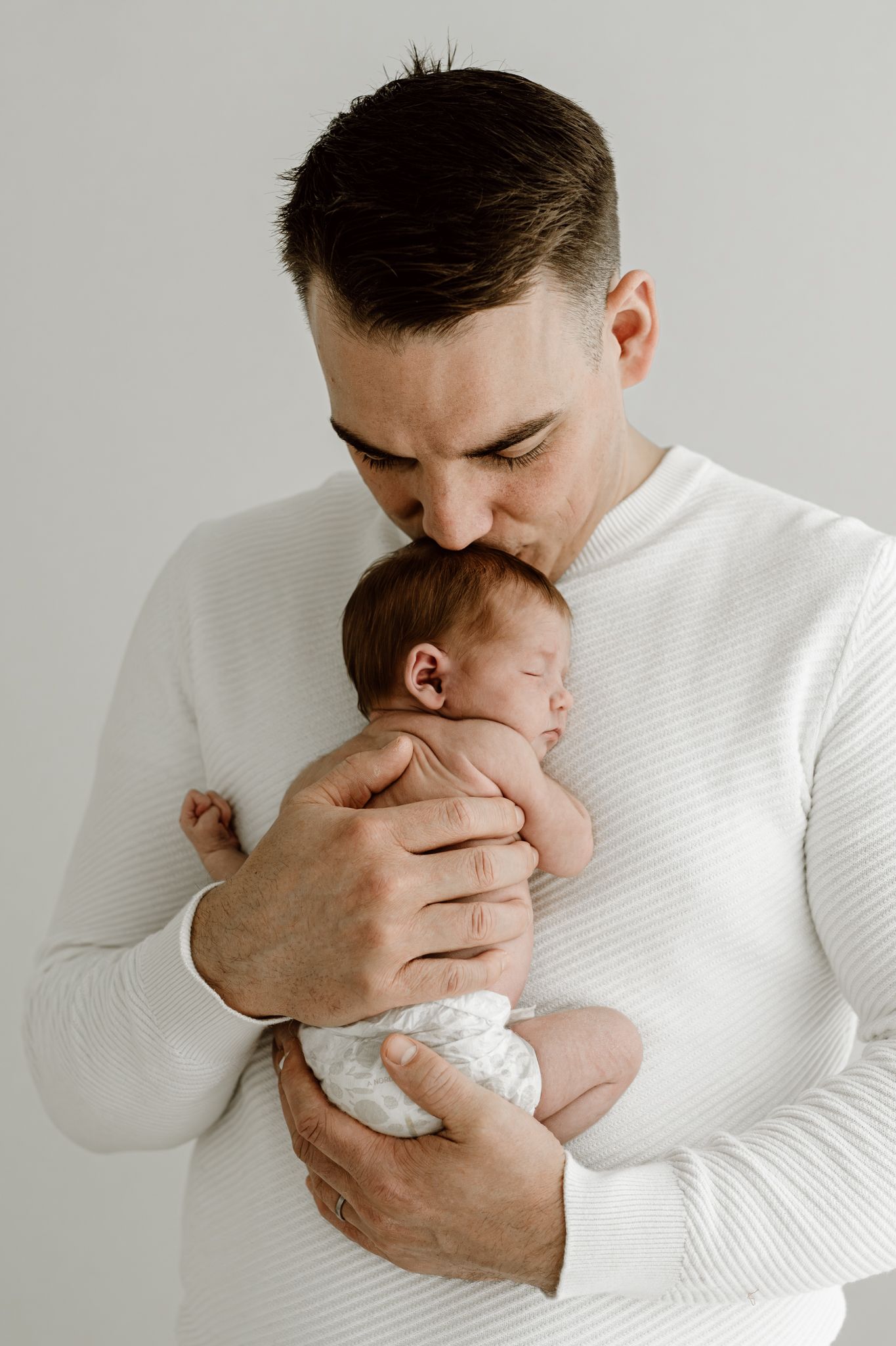 Father in white ribbed sweater gently kisses sleeping newborn baby he holds.