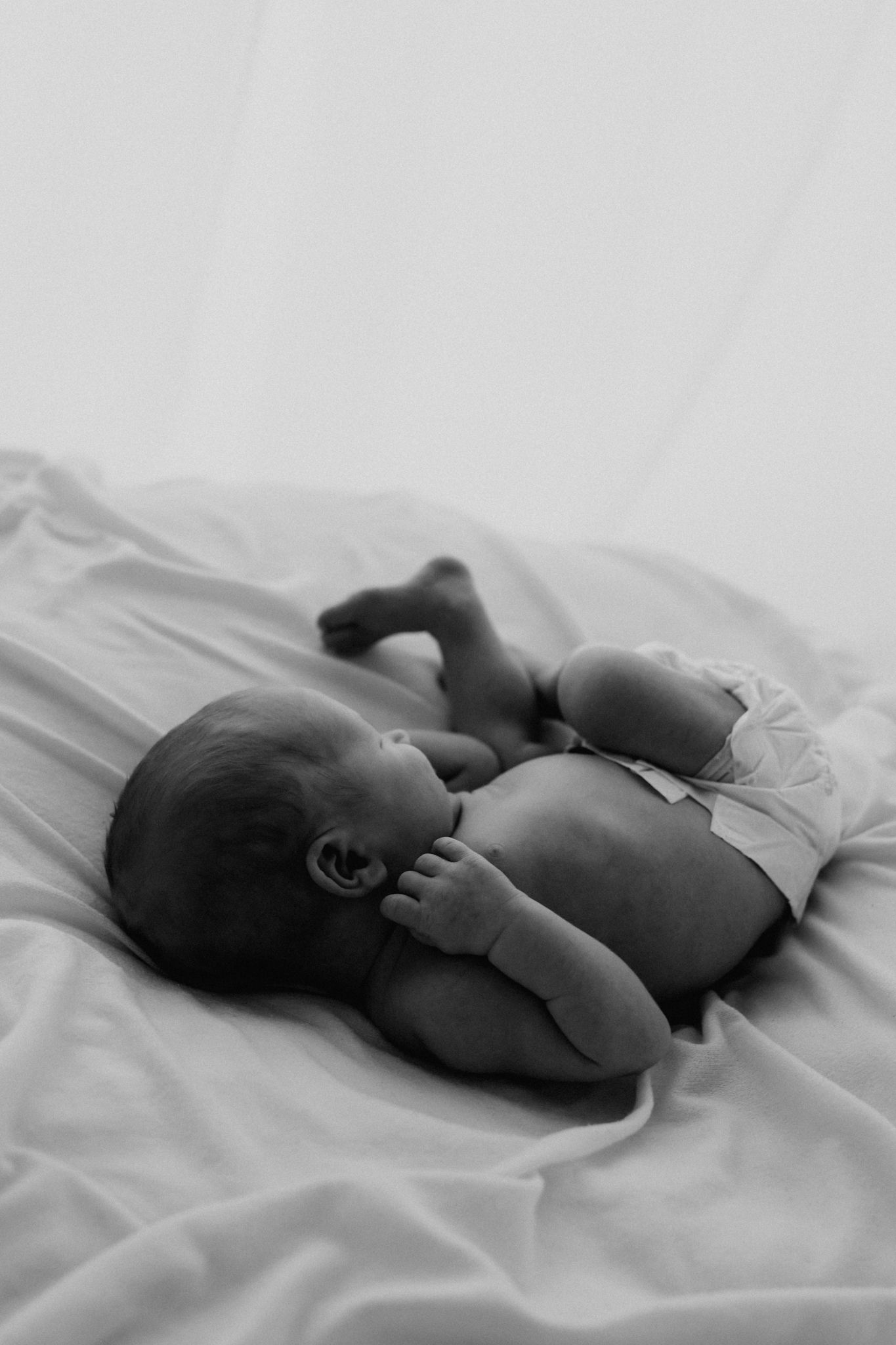 Black and white photo of a newborn baby lying on its side on soft white fabric in fetal position.