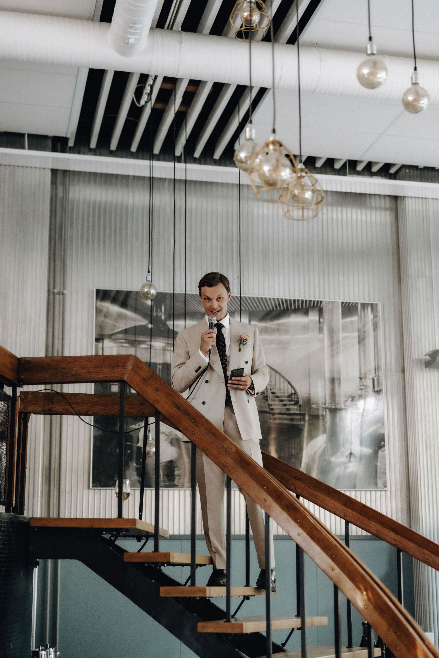 Groom in cream suit descending spiral staircase holding microphone at wedding reception.