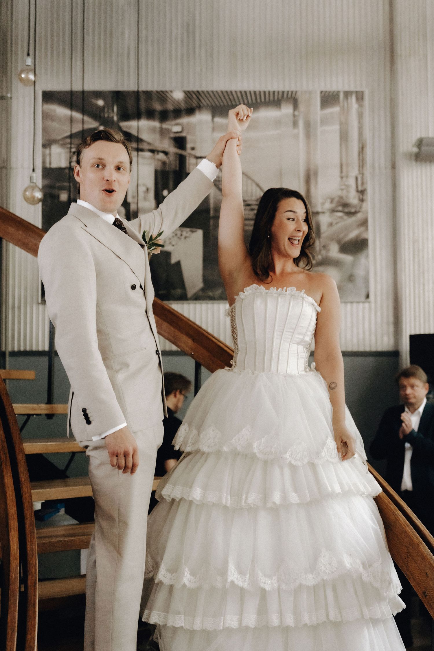 Joyful newlyweds raise hands in celebration on staircase at their indoor wedding reception venue.