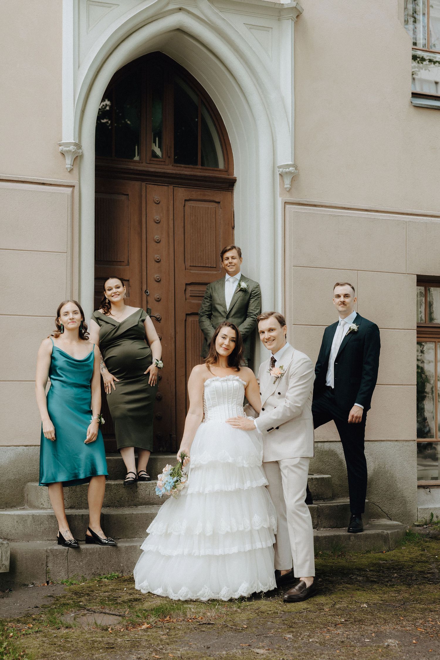 Wedding group portrait outside church entrance, bride in layered white dress embraced by groom in beige suit.