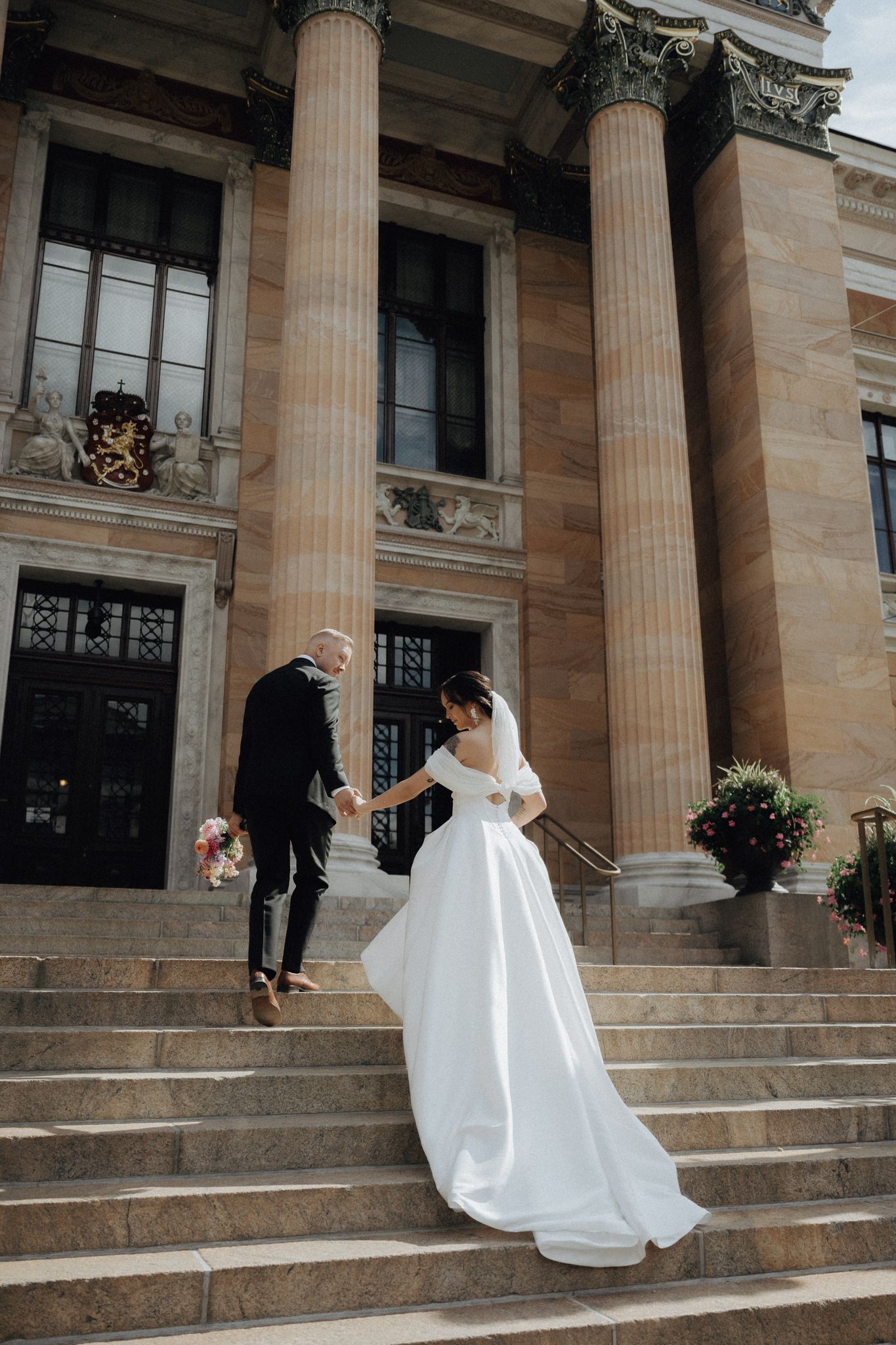Bride and groom holding hands on grand stone steps of a classical building with tall columns.