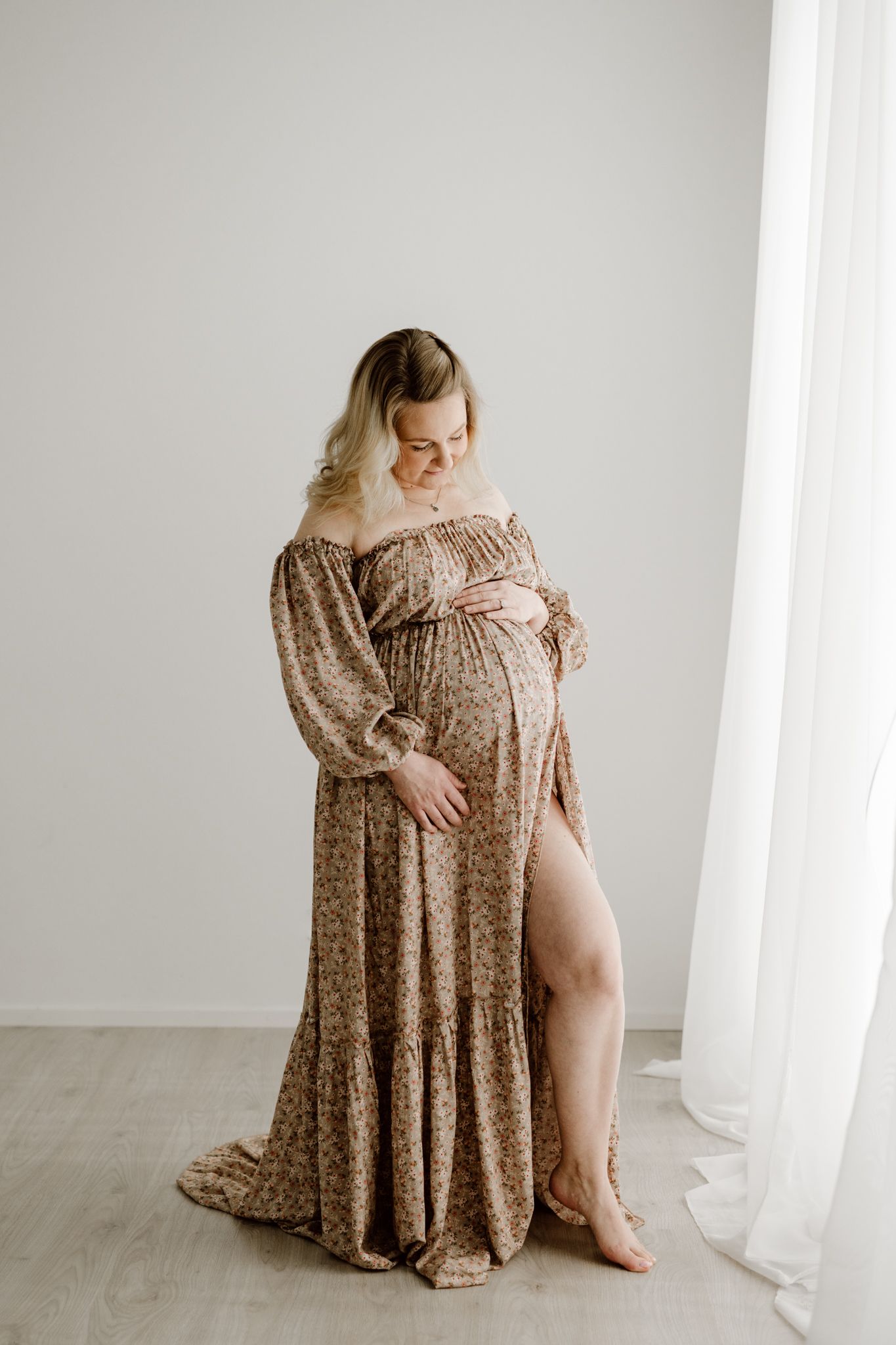 Pregnant woman in flowing floral off-shoulder gown with leg slit, cradling bump near sheer curtain in studio.