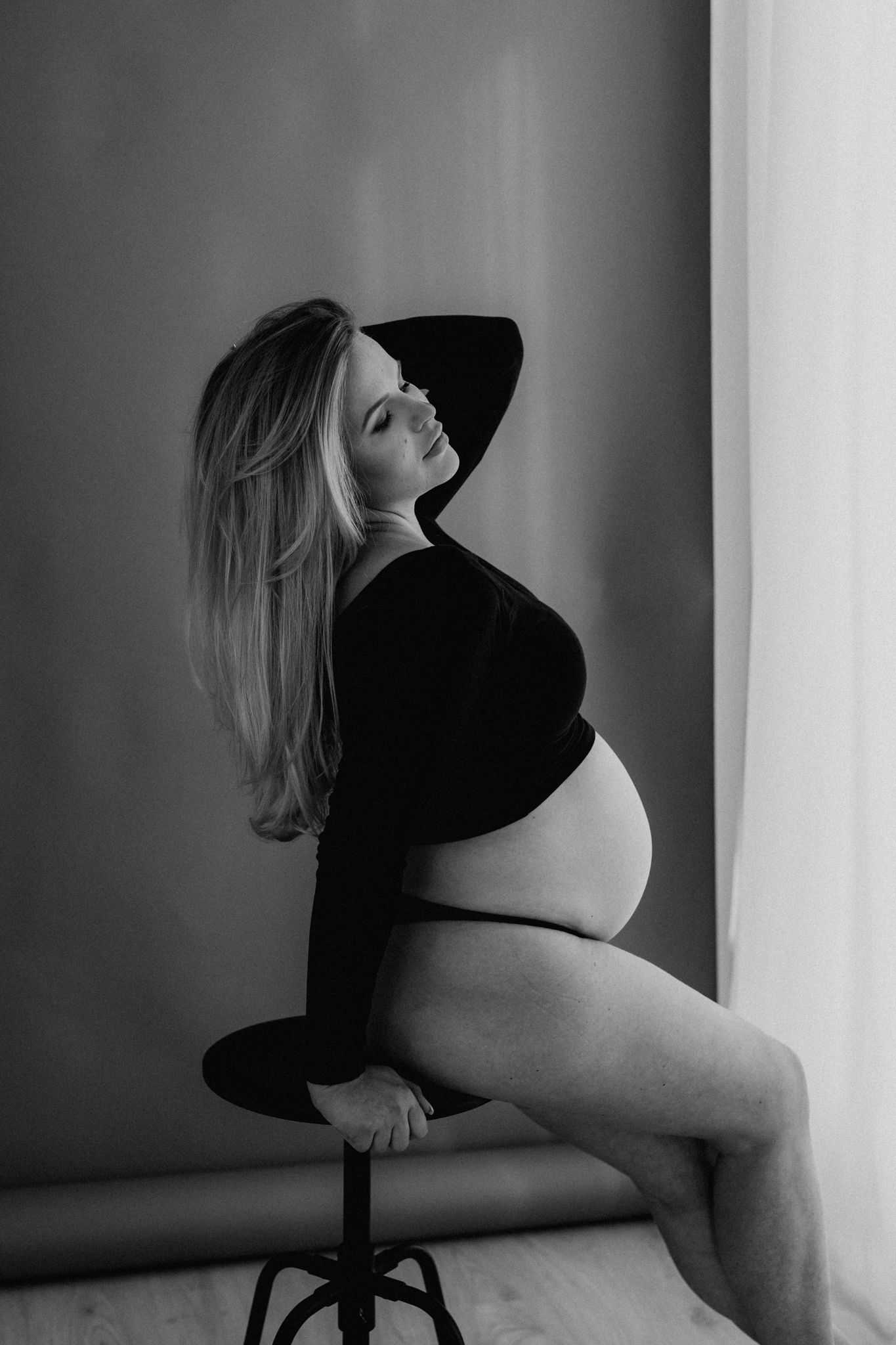 Black and white side profile of pregnant woman in black crop top seated on stool.