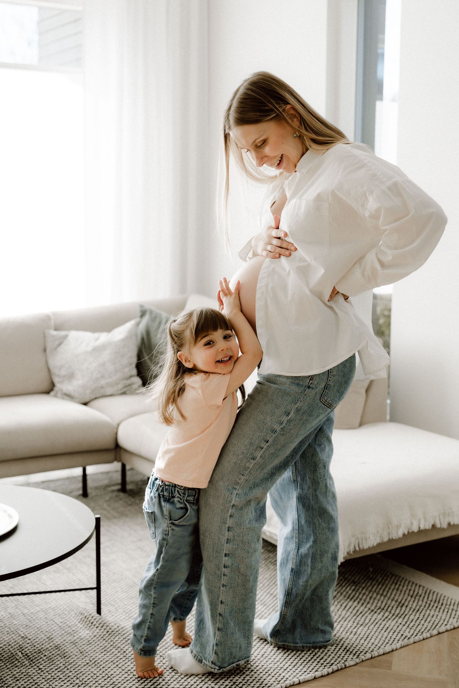 Pregnant woman in white shirt and jeans smiling down at young daughter hugging her baby bump in bright living room.