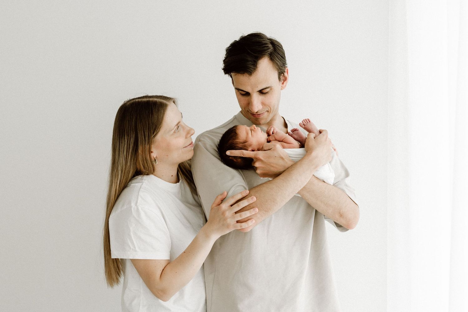 Young parents in white shirts holding and admiring their swaddled newborn baby.